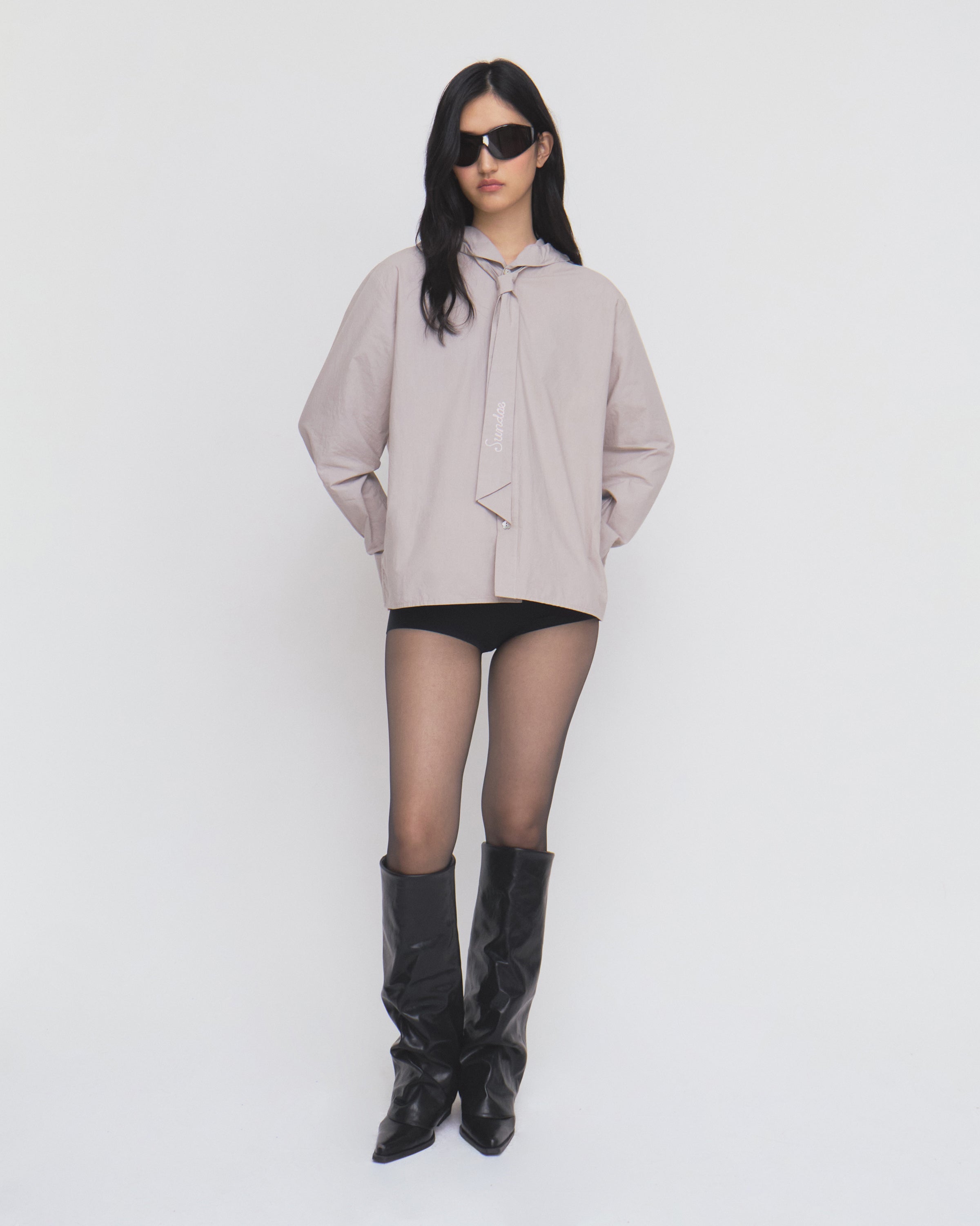 Model in black sunglasses, the Sundae School GREY SUNBAE TIE SHIRT, black shorts, sheer black tights, and black knee-high boots stands against a plain white background.