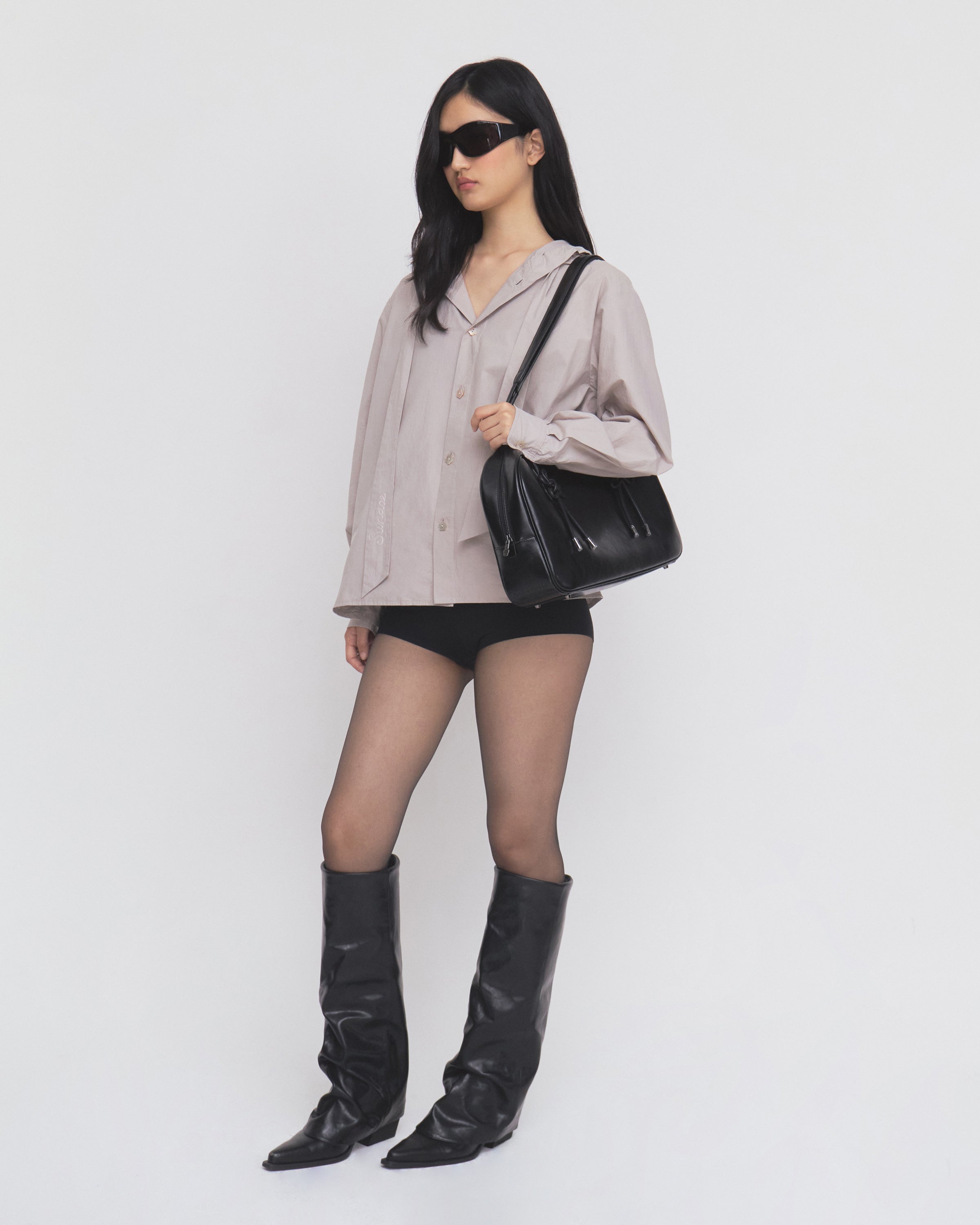 A woman wearing the GREY SUNBAE TIE SHIRT by Sundae School, styled with sunglasses, black shorts, sheer tights, black knee-high boots, and carrying a black handbag stands against a plain white background.