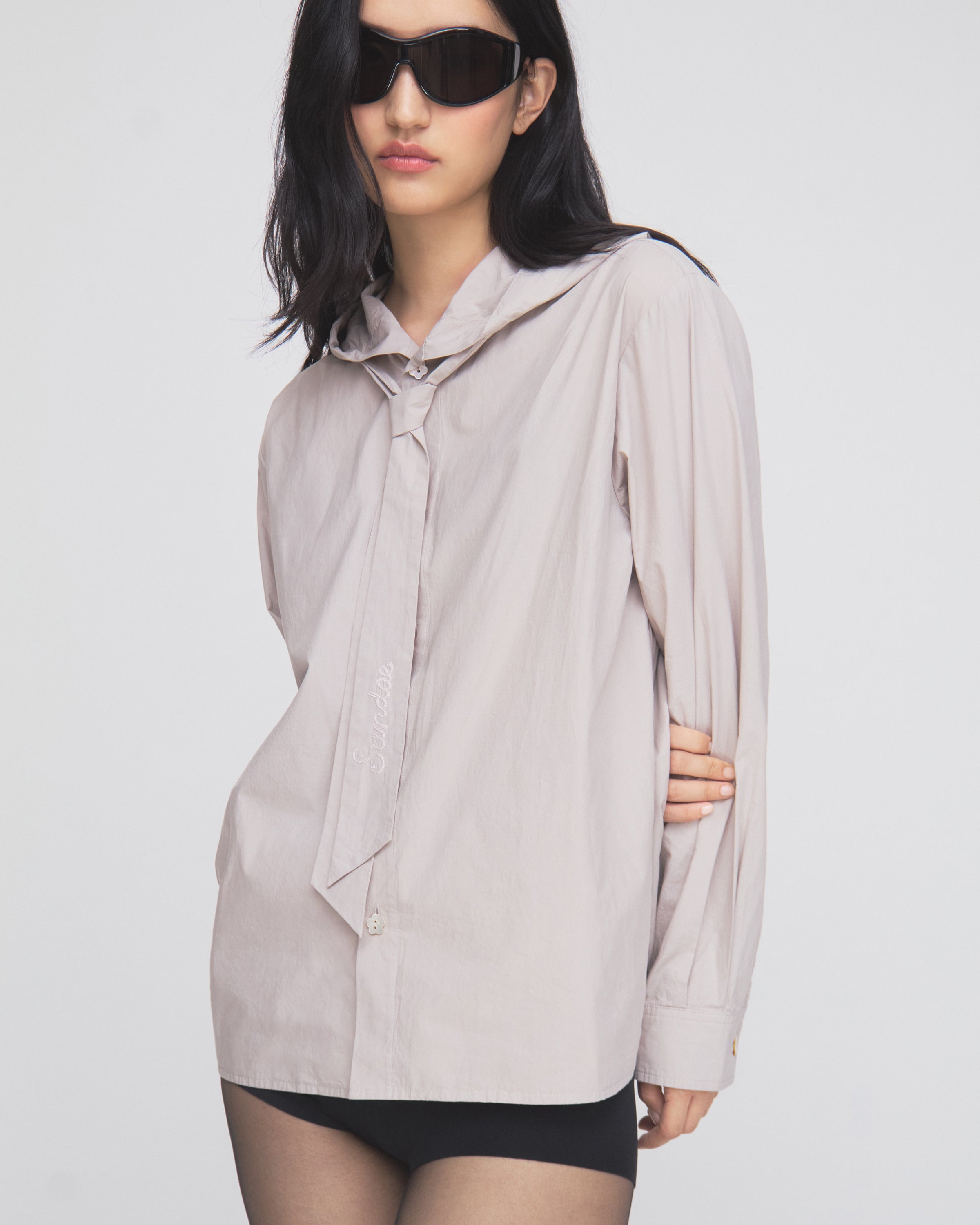 A woman with long dark hair wears black sunglasses and the Sundae School GREY SUNBAE TIE SHIRT—a light-colored button-up shirt with a loose necktie—paired with black shorts, standing against a plain white background.