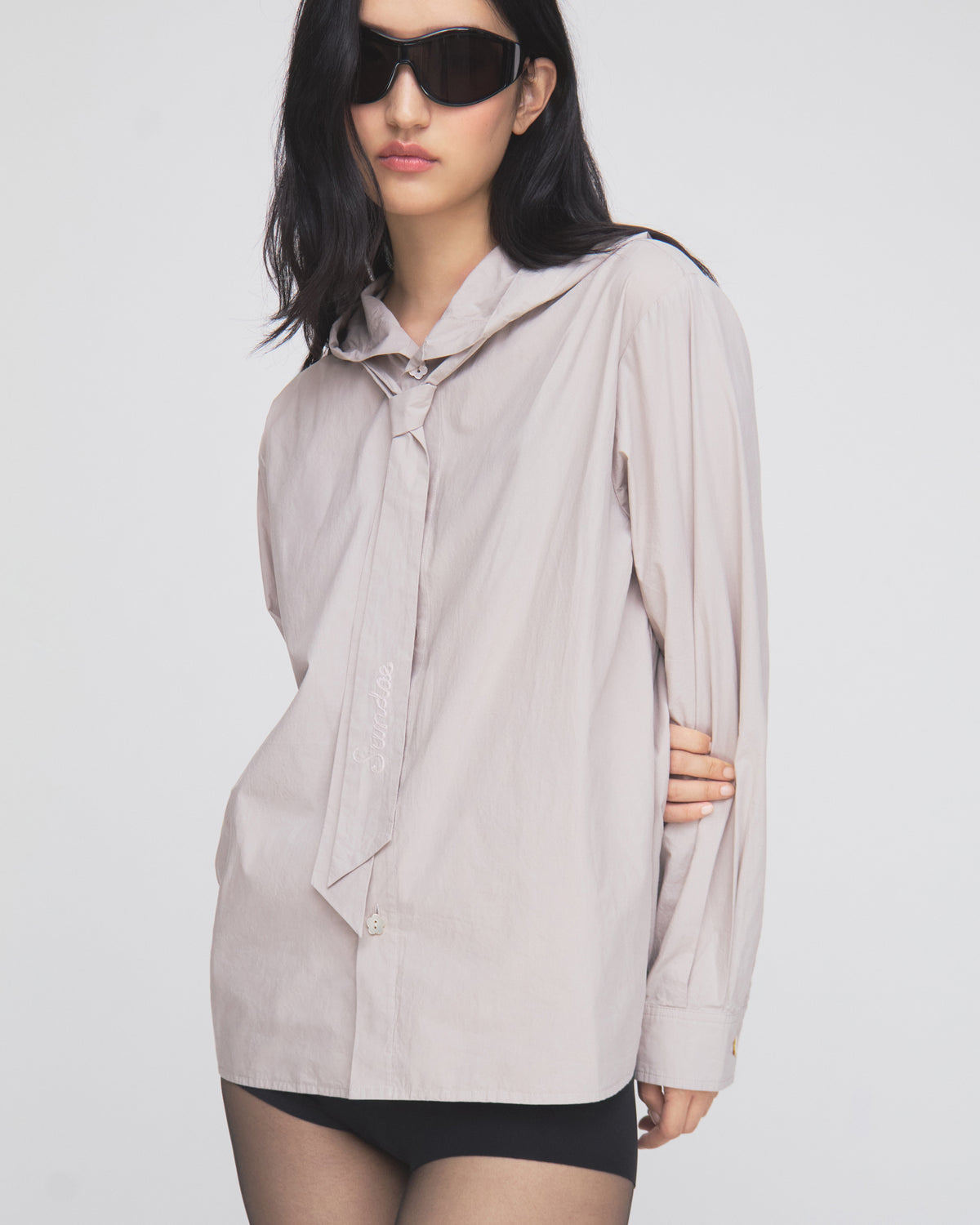 A woman with long dark hair wears black sunglasses and the Sundae School GREY SUNBAE TIE SHIRT—a light-colored button-up shirt with a loose necktie—paired with black shorts, standing against a plain white background.