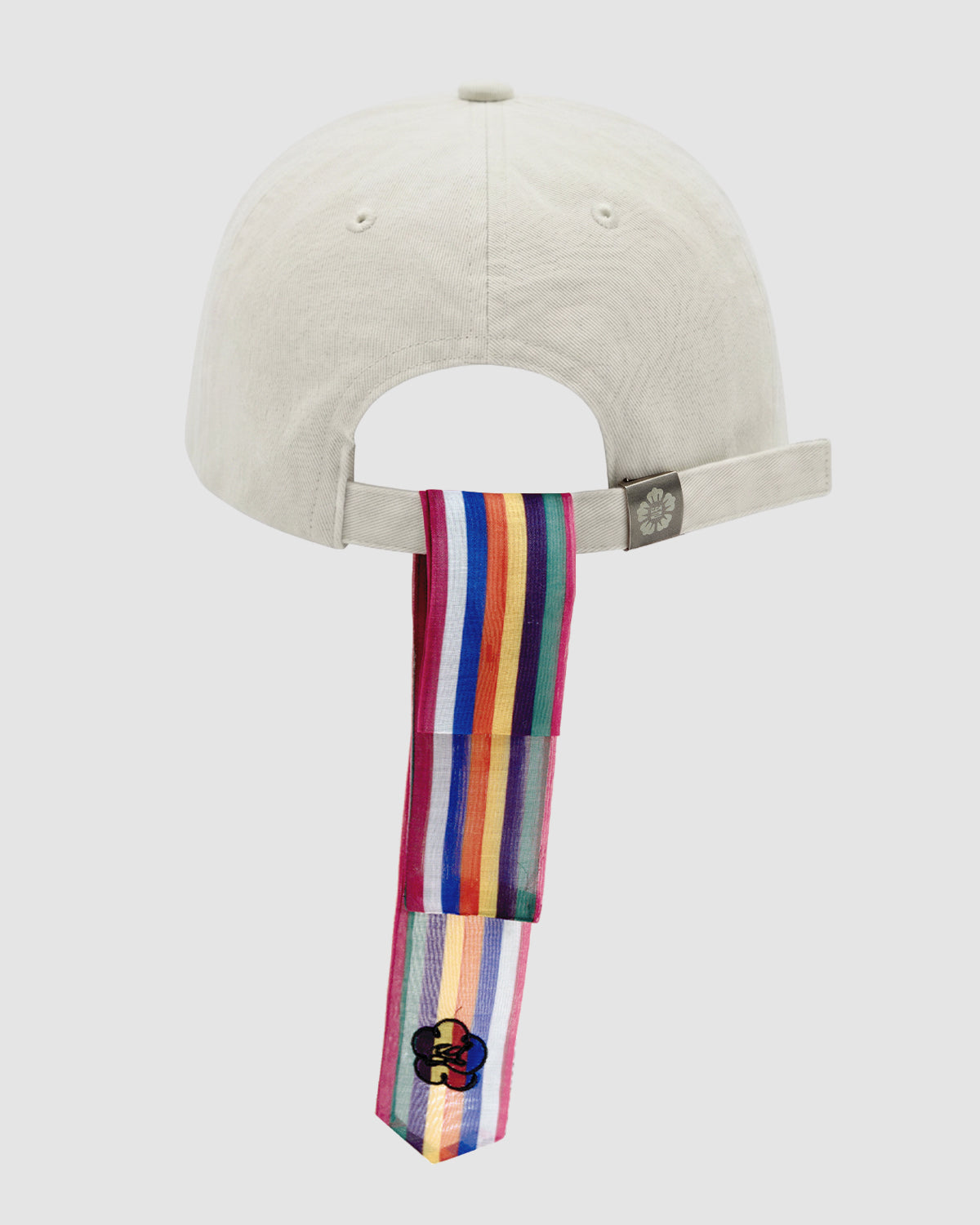 The Sundae School IVORY DAENGGI CAP is a light-colored adjustable cotton cap seen from the back, with a rainbow-striped daenggi attached to the strap and hanging below, featuring a flower patch at the end for a playful accent.