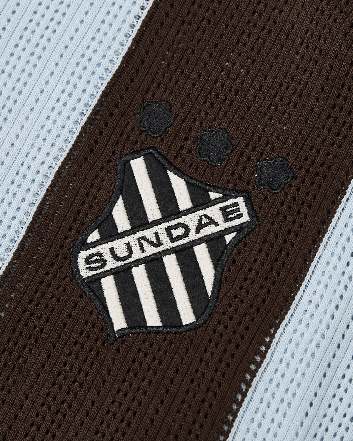 Close-up of a SKY BLUE SUNDAE SOCCER JERSEY by Sundae School, featuring light blue and dark brown striped cotton knit, with a central embroidered chest patch that reads 