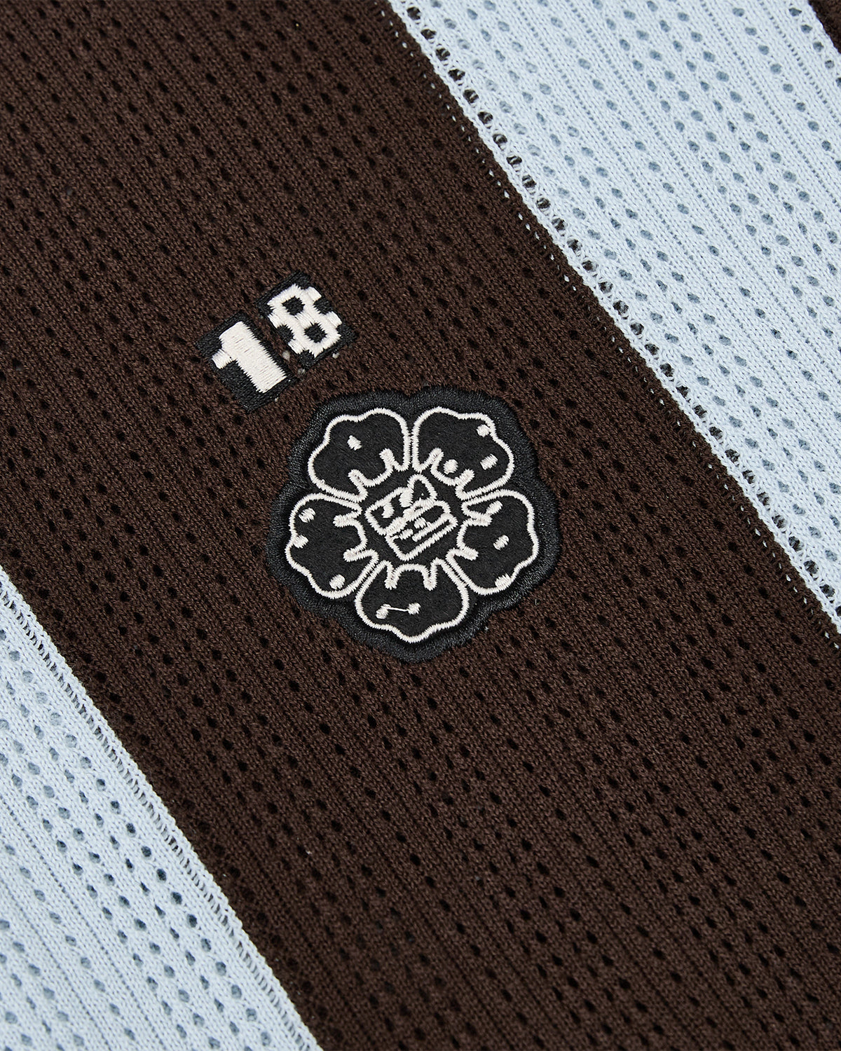 Close-up of the SKY BLUE SUNDAE SOCCER JERSEY by Sundae School, featuring lightweight cotton knit with brown and light blue stripes, the number 18, and a central black and white floral emblem inspired by mochi buttons.