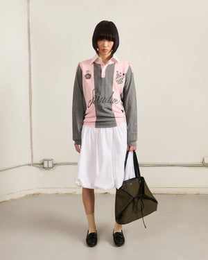 A person with a short bob wears the Sundae School PINK SUNDAE SOCCER JERSEY, a 100% cotton lightweight top, paired with a white skirt, beige socks, black shoes, and holding an olive green bag in a plain room with exposed outlets and cords.