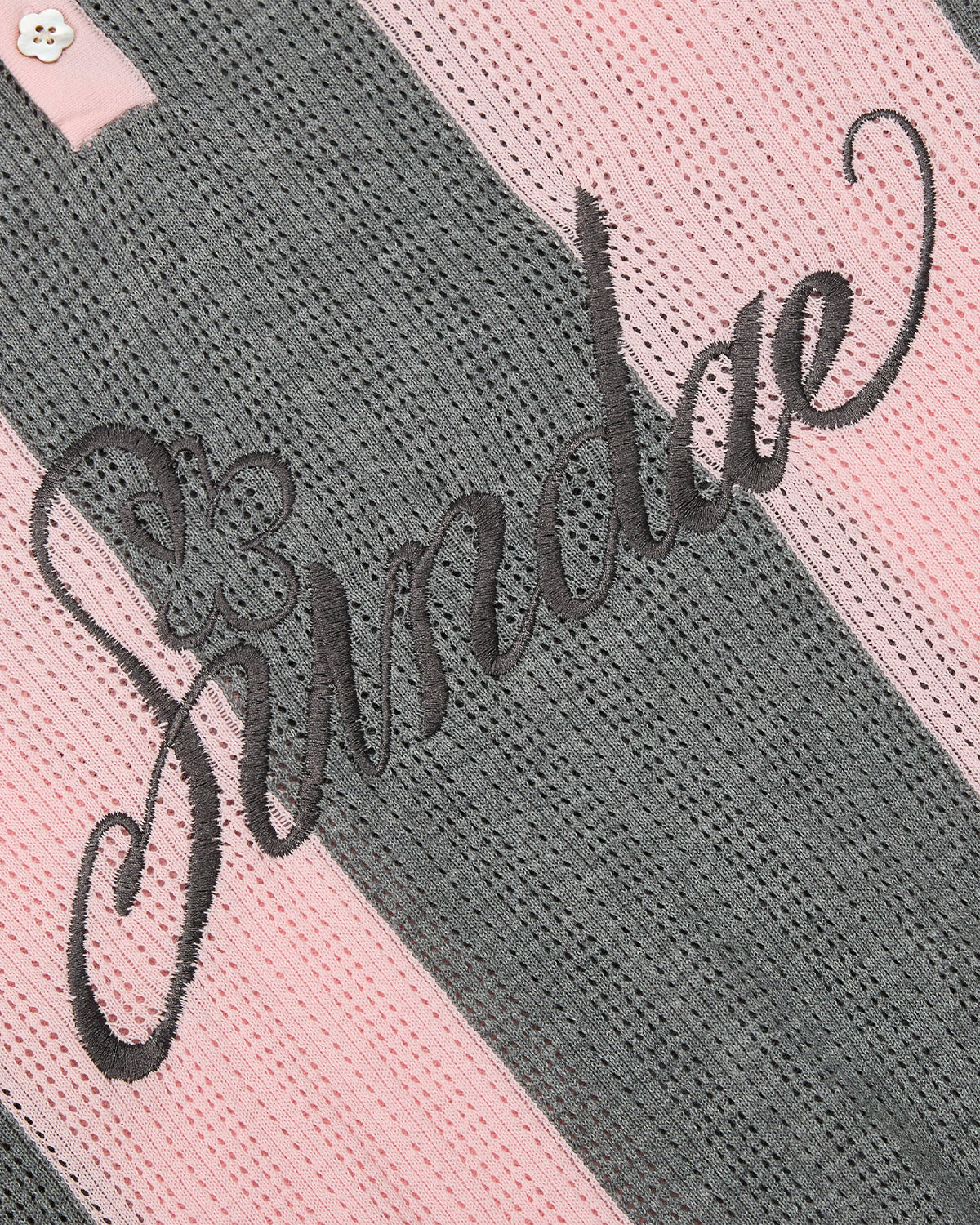 A close-up of the PINK SUNDAE SOCCER JERSEY by Sundae School reveals its pink and gray striped perforated knit fabric, highlighted with “Sundae” embroidered in black cursive and a delicate floral accent. Crafted from 100% cotton, this lightweight top features custom embroidered chest patches, adding a unique and stylish touch.