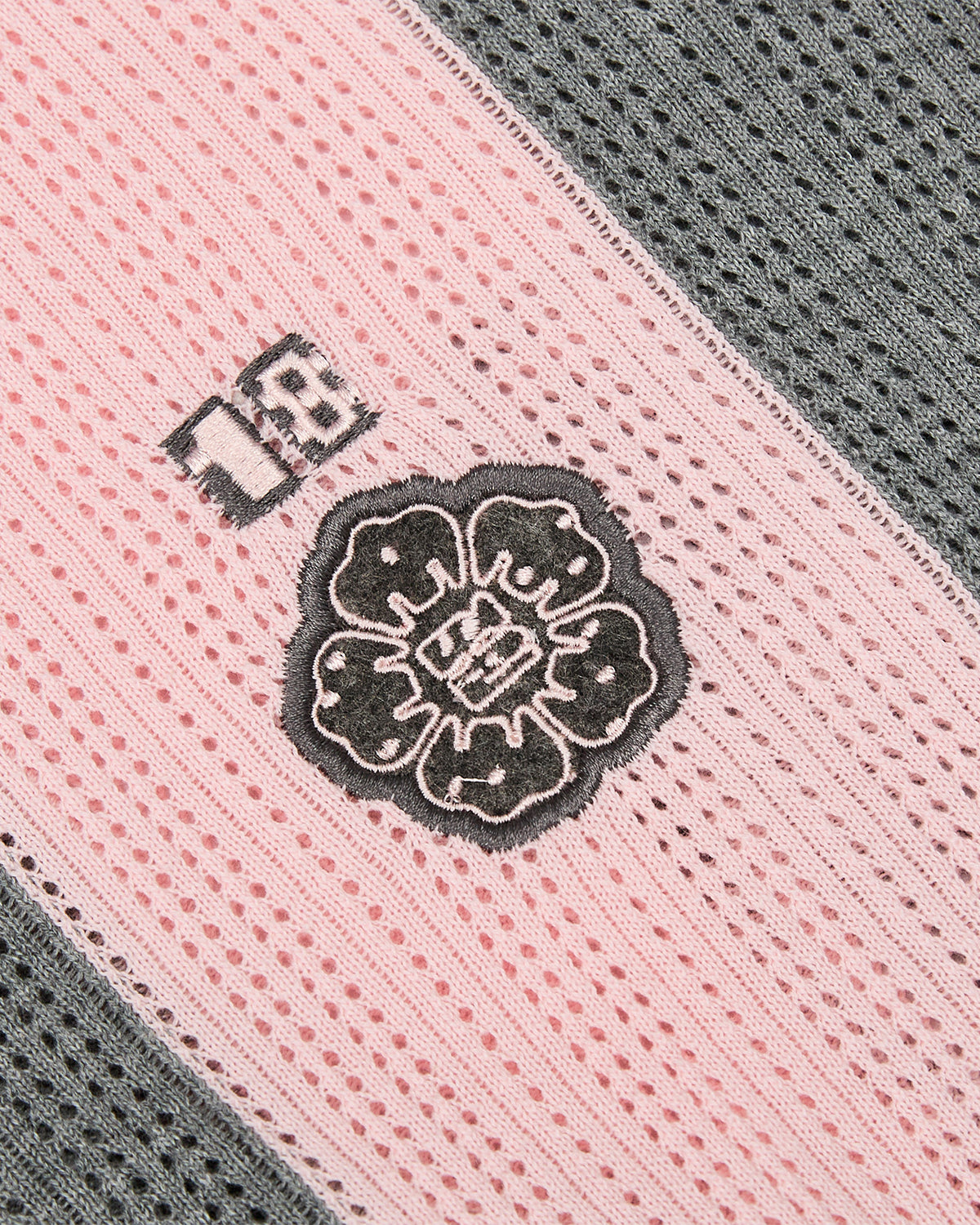 Close-up of the Sundae School PINK SUNDAE SOCCER JERSEY, showcasing pink and gray lace fabric with a black embroidered flower detail and a small custom chest patch featuring the number 18 on the pink section. The jersey’s lace displays a repeating circular, perforated knit pattern that adds textured dimension to this delicate design.