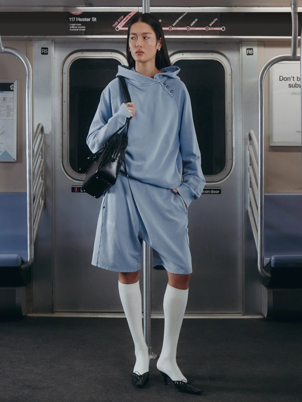 A model stands on a subway train wearing a light blue oversized hooded dress with a long tie at the waist, white knee-high socks, black shoes, and carrying a black shoulder bag.