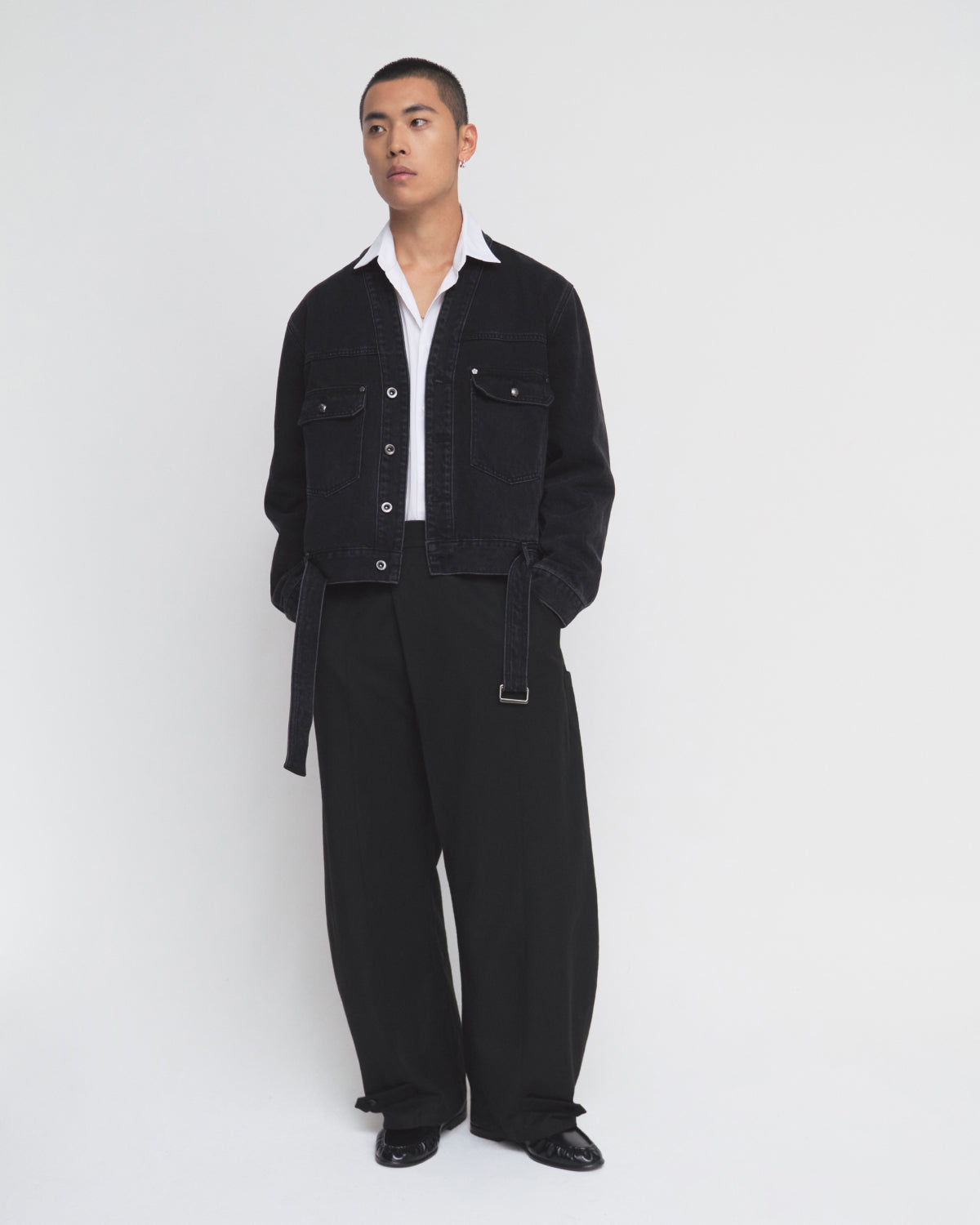 A person stands against a plain white background wearing the BLACK WIDOW TAEKWONDO DENIM JACKET by Sundae School over a white collared shirt and loose black pants with a double-buckle belt and black shoes, looking to the side with hands in pockets.