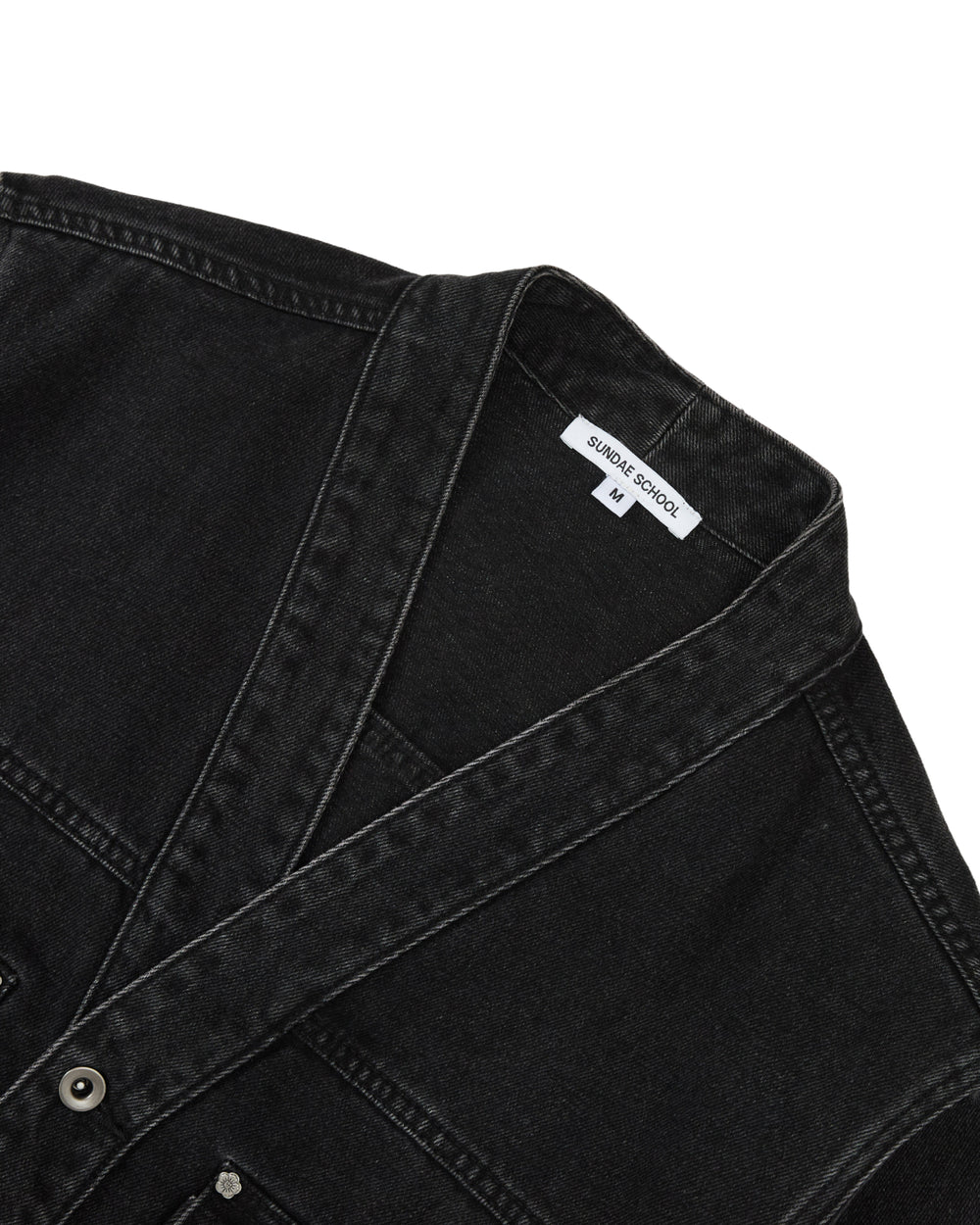Close-up of the BLACK WIDOW TAEKWONDO DENIM JACKET by Sundae School, featuring visible stitching, metal buttons, and a white collar tag that reads “Sundae School” and “M.” Reminiscent of a modern Korean Canadian tuxedo, the black denim jacket is laid flat against a white background.