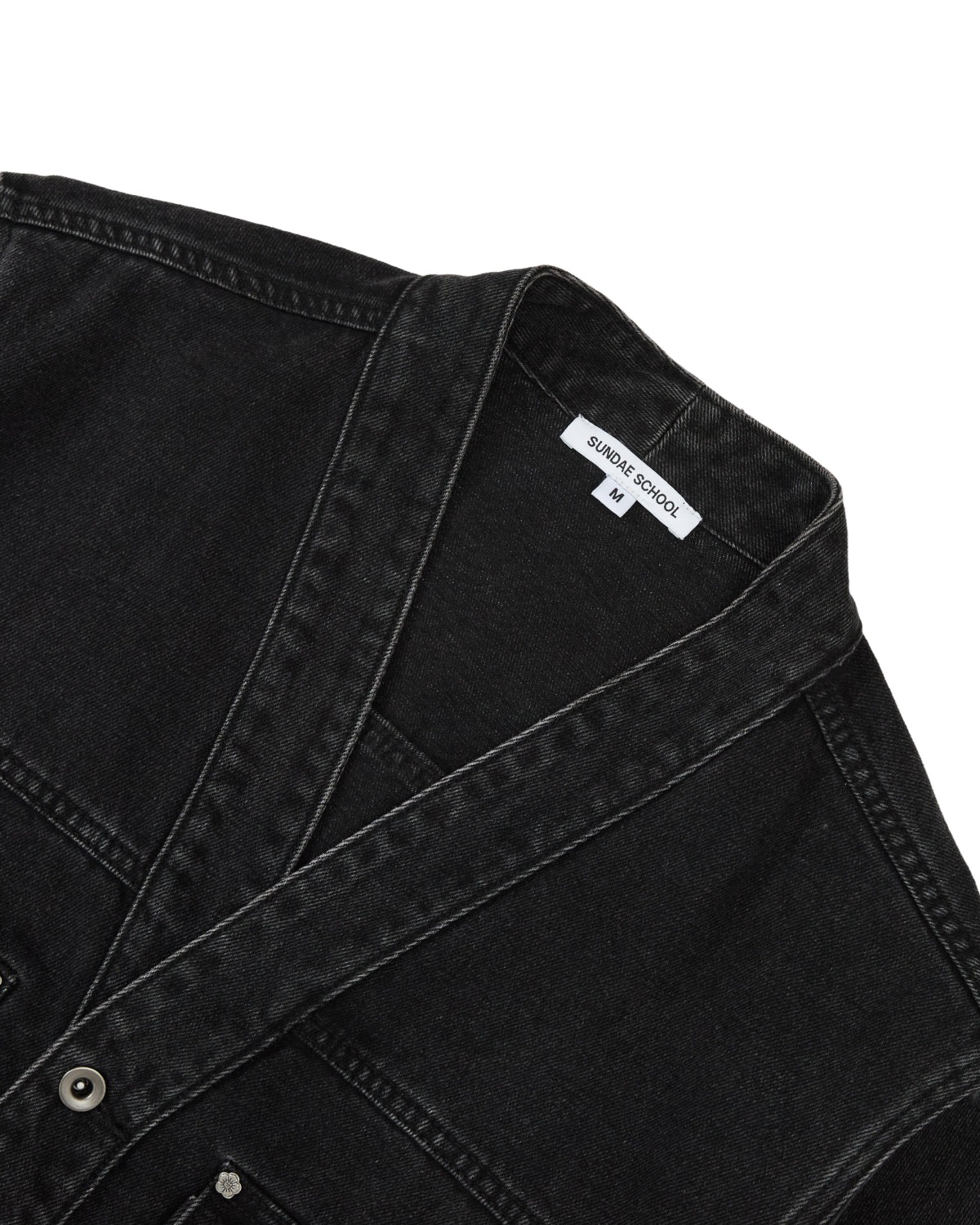 Close-up of the BLACK WIDOW TAEKWONDO DENIM JACKET by Sundae School, featuring visible stitching, metal buttons, and a white collar tag that reads “Sundae School” and “M.” Reminiscent of a modern Korean Canadian tuxedo, the black denim jacket is laid flat against a white background.