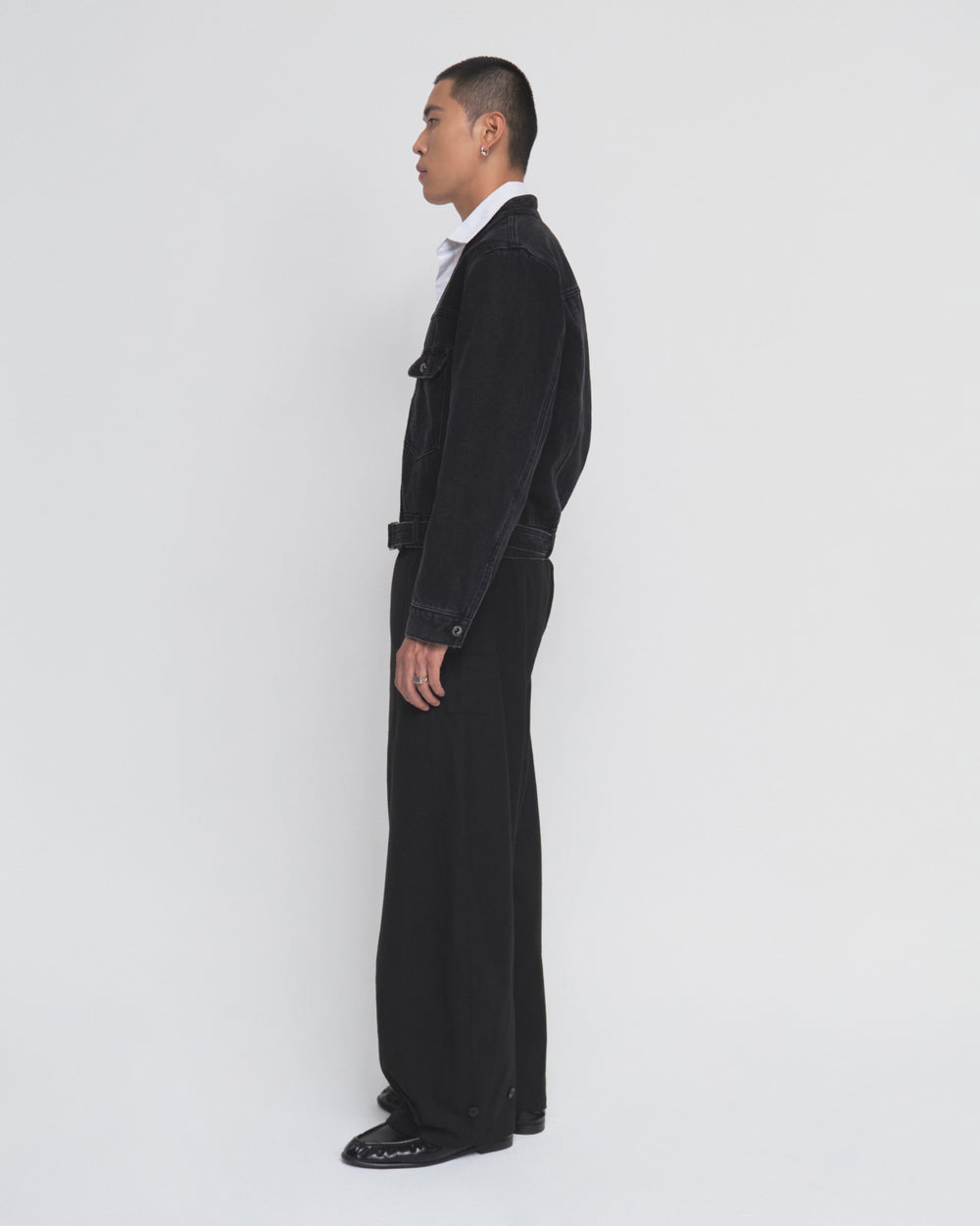 A person stands in profile against a plain white background, wearing the BLACK WIDOW TAEKWONDO DENIM JACKET by Sundae School, styled as part of a modern Korean Canadian tuxedo with wide-leg black pants and black shoes.