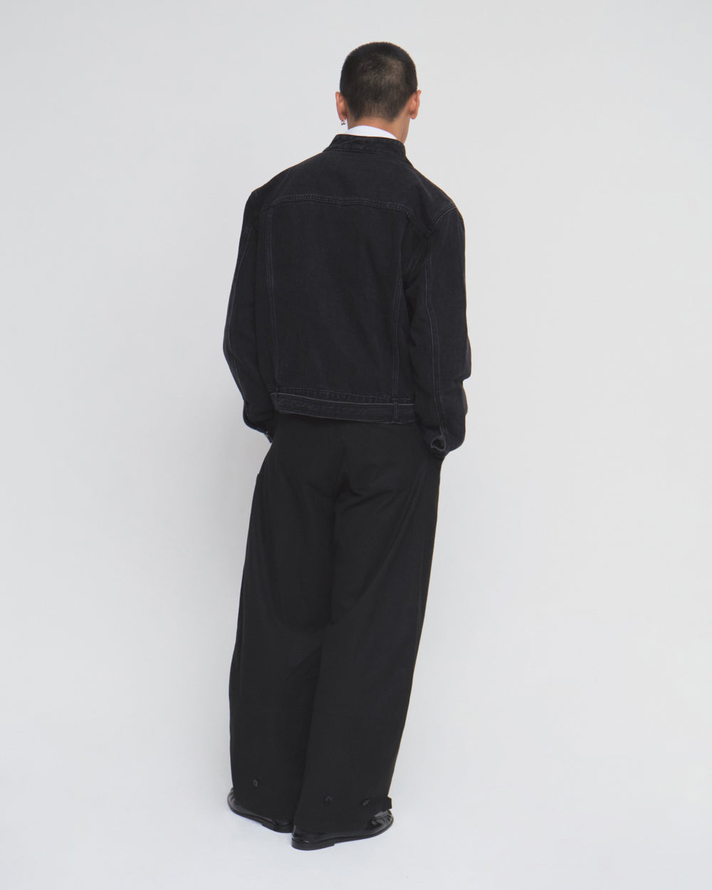 A person stands facing away, wearing the Sundae School BLACK WIDOW TAEKWONDO DENIM JACKET and wide-leg black pants with a double-buckle belt, set against a plain white background.