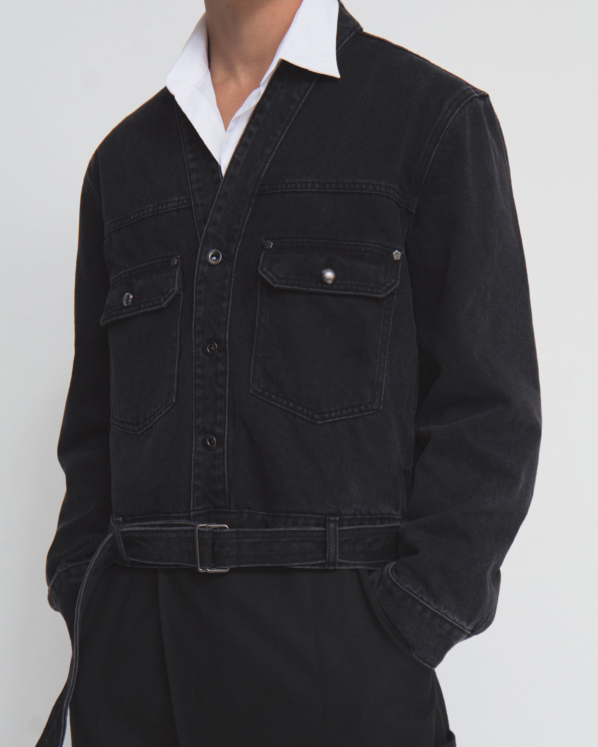 A person wearing the BLACK WIDOW TAEKWONDO DENIM JACKET by Sundae School—a black Korean Canadian tuxedo-style denim jacket with chest pockets and a double-buckle belt—layered over a white collared shirt and black pants, stands against a plain light background.