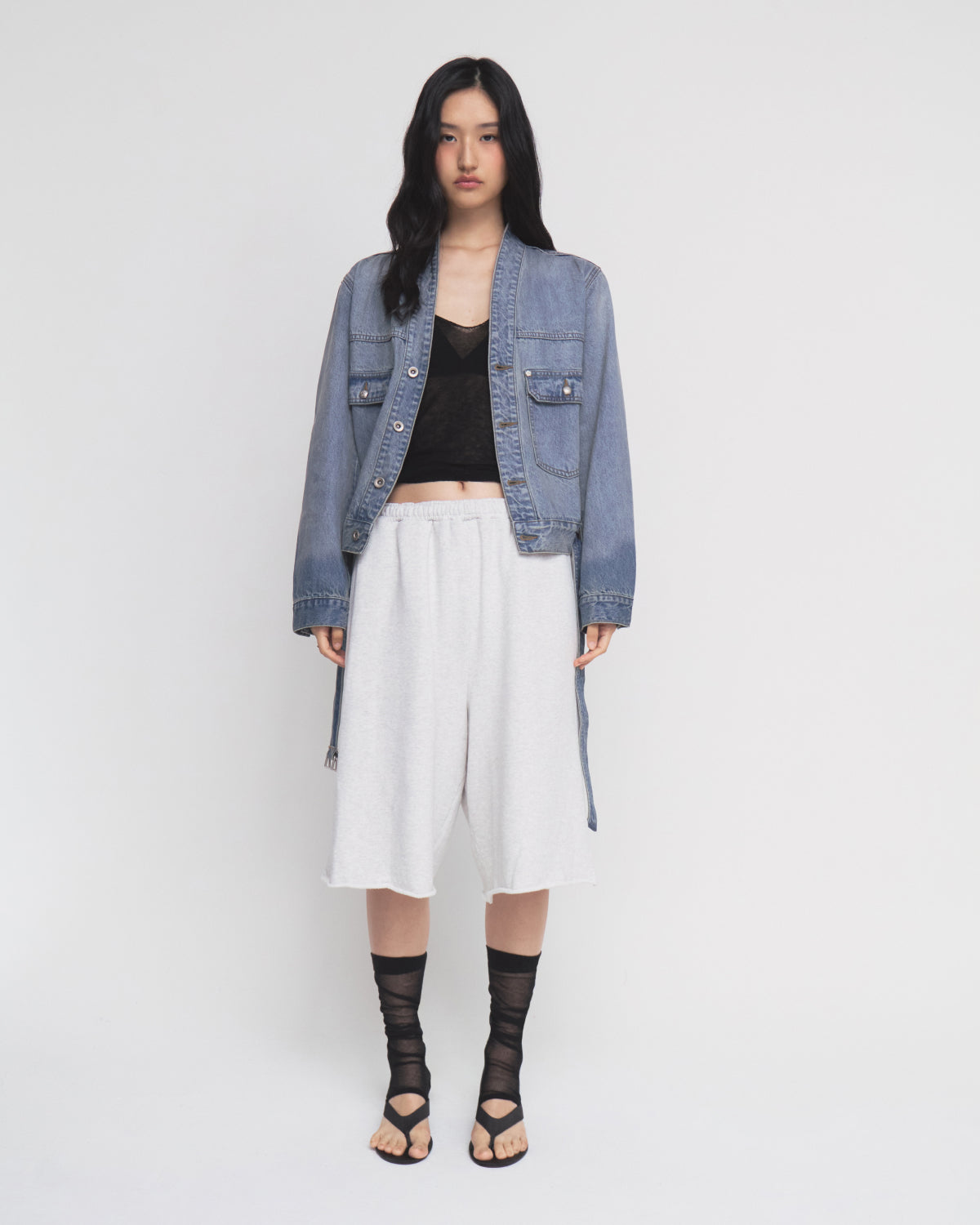 A woman stands against a white background wearing the MEDIUM BLUE TAEKWONDO DENIM JACKET by Sundae School, paired with a black top, loose white shorts with a detachable belt, and black strappy sandals with sheer black socks.