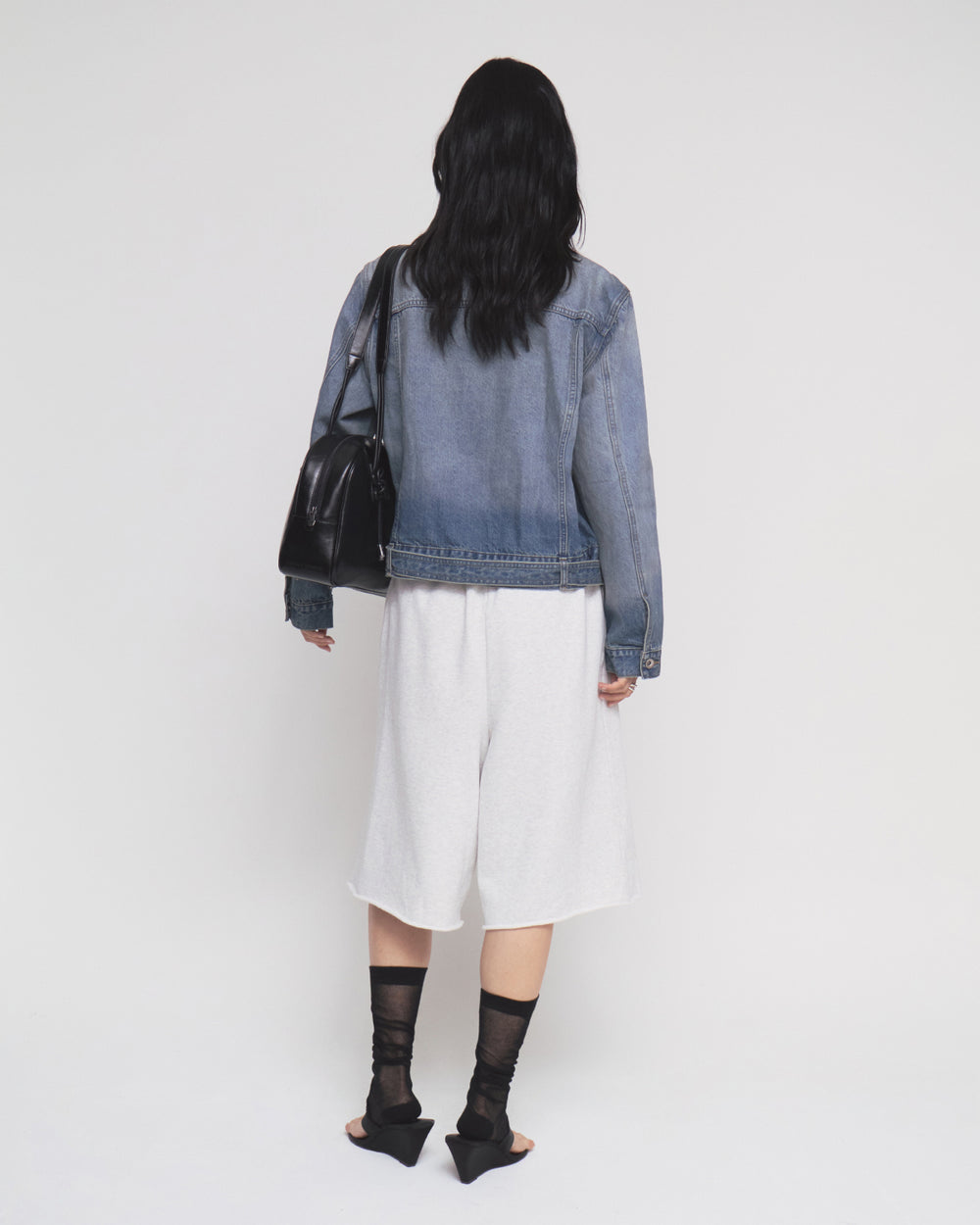 A person with long dark hair is seen from behind, wearing the Sundae School MEDIUM BLUE TAEKWONDO DENIM JACKET styled as a Korean Canadian tuxedo, paired with white knee-length shorts, black sheer socks, and black heeled sandals. They carry a black shoulder bag with a detachable belt. The background is plain white.