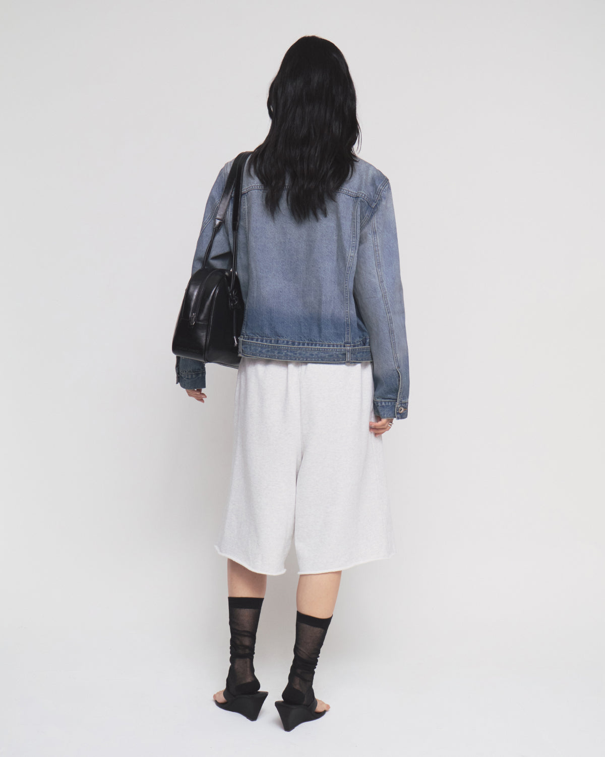 A person with long dark hair is seen from behind, wearing the Sundae School MEDIUM BLUE TAEKWONDO DENIM JACKET styled as a Korean Canadian tuxedo, paired with white knee-length shorts, black sheer socks, and black heeled sandals. They carry a black shoulder bag with a detachable belt. The background is plain white.