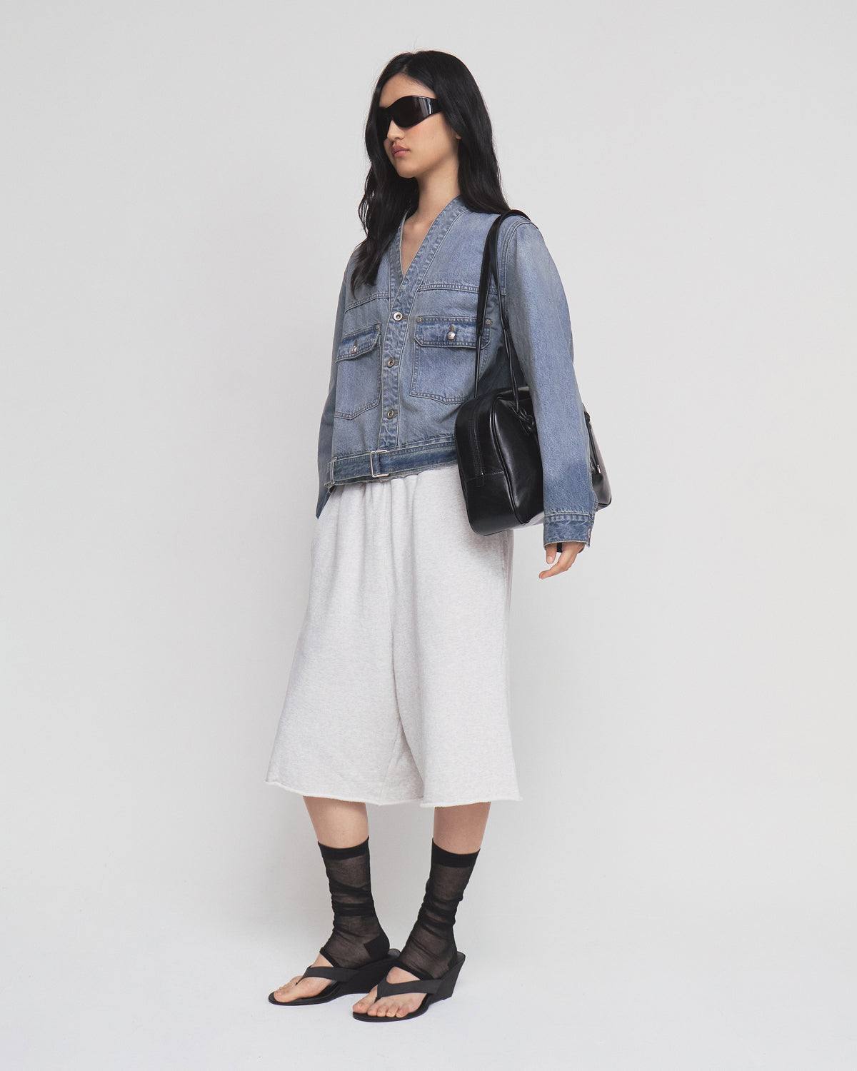 A woman wearing sunglasses, the MEDIUM BLUE TAEKWONDO DENIM JACKET by Sundae School styled in a Korean Canadian tuxedo fashion, a white skirt, black sheer socks, black sandals, and carrying a black handbag stands against a plain white background.
