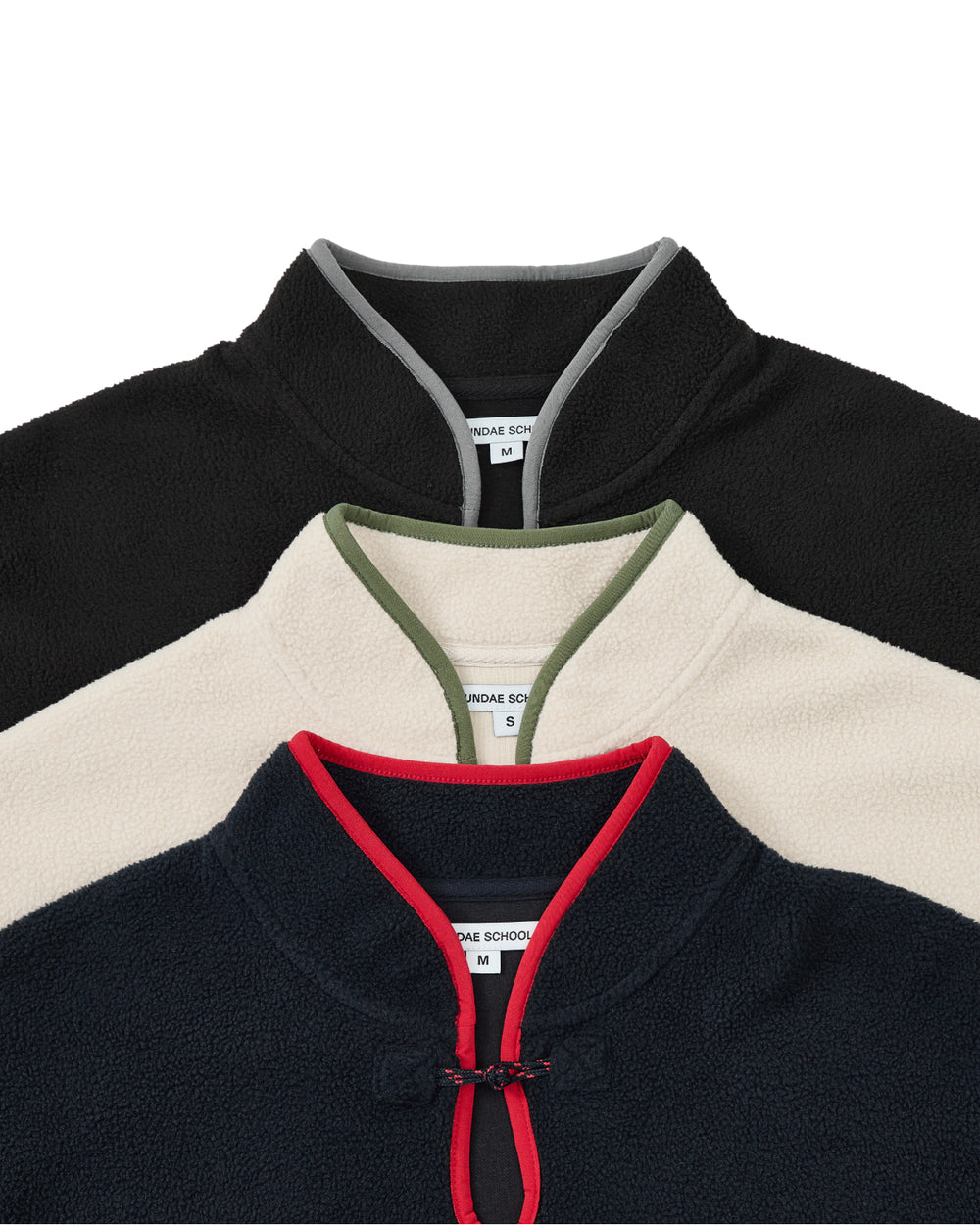 Three cozy Sundae School NAVY TEARDROP KNOT FLEECE jackets are stacked. The top jacket is black with a gray trim, the middle one is off-white with an olive trim, and the bottom features a navy body with a red trim. Each has a stand collar and visible Medium size tags, all showcasing an East Asian–inspired fleece aesthetic that effortlessly blends comfort and style.