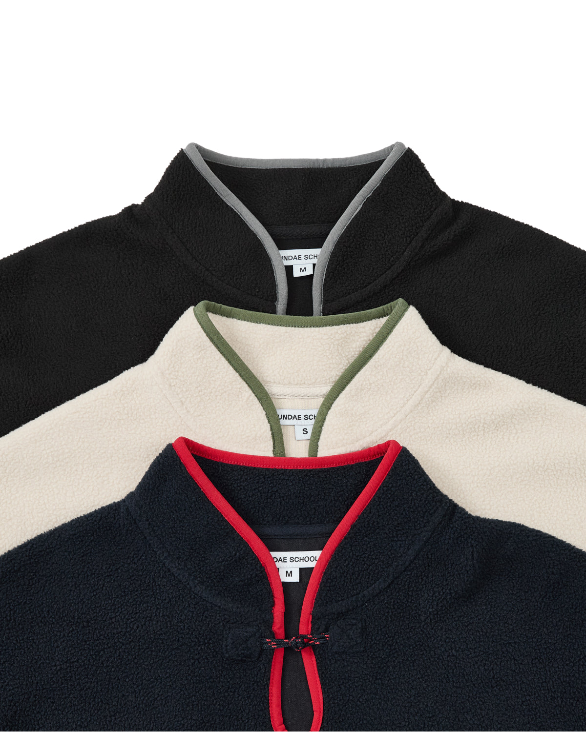Three cozy Sundae School NAVY TEARDROP KNOT FLEECE jackets are stacked. The top jacket is black with a gray trim, the middle one is off-white with an olive trim, and the bottom features a navy body with a red trim. Each has a stand collar and visible Medium size tags, all showcasing an East Asian–inspired fleece aesthetic that effortlessly blends comfort and style.