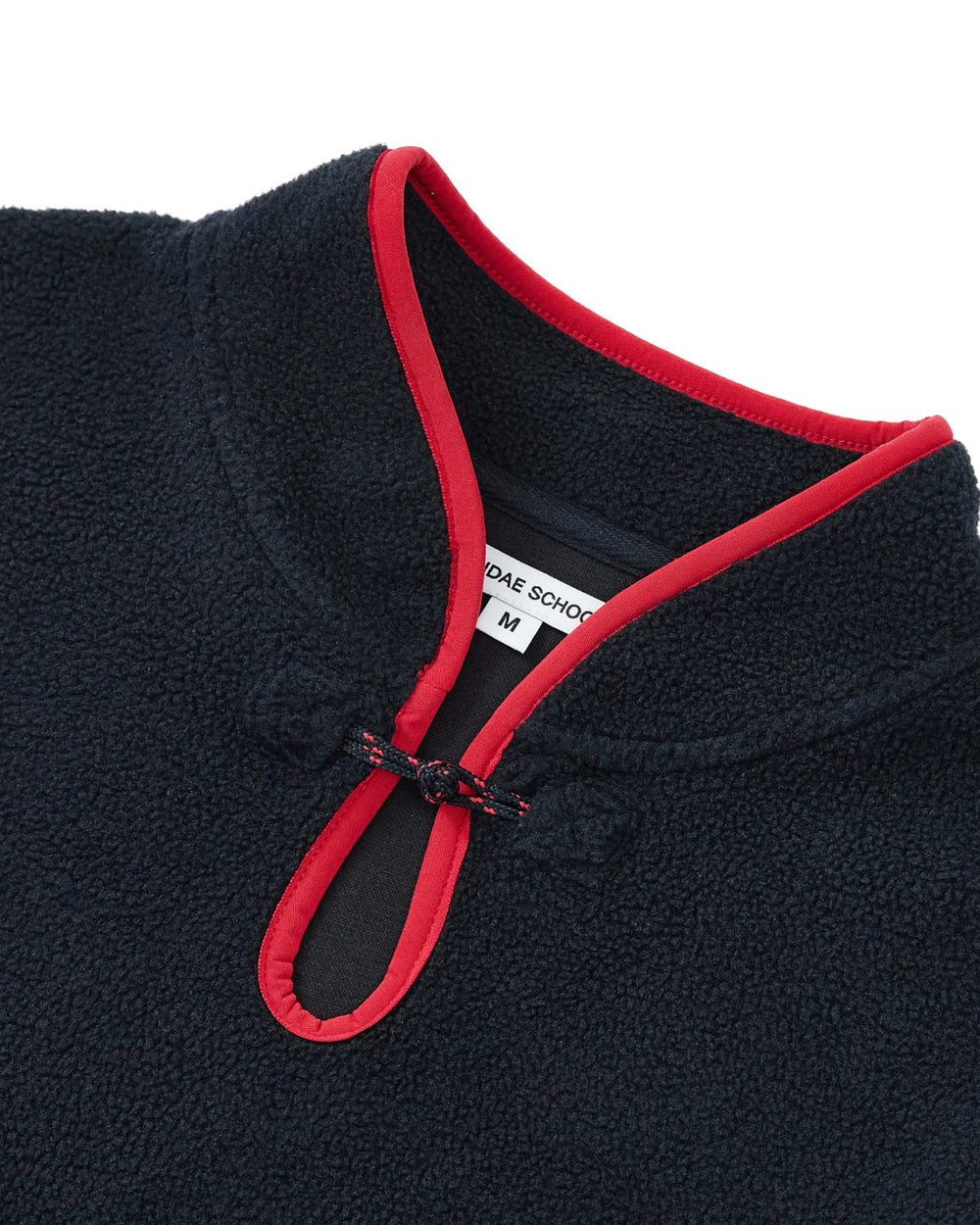 Close-up of the NAVY TEARDROP KNOT FLEECE by Sundae School, featuring a cozy navy blue fleece with red trim around the collar and keyhole opening, secured by an East Asian inspired teardrop knot. A white inner tag displays the size M along with a partial school name.