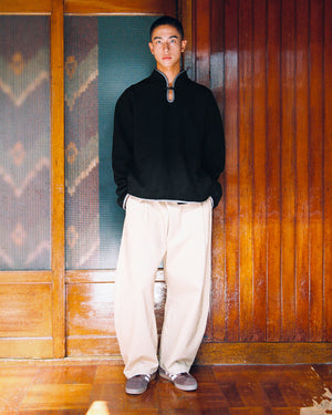 A person stands indoors against a wooden wall, wearing the Sundae School BLACK TEARDROP KNOT FLEECE, wide beige pants, and gray shoes, with hands in pockets, looking at the camera.