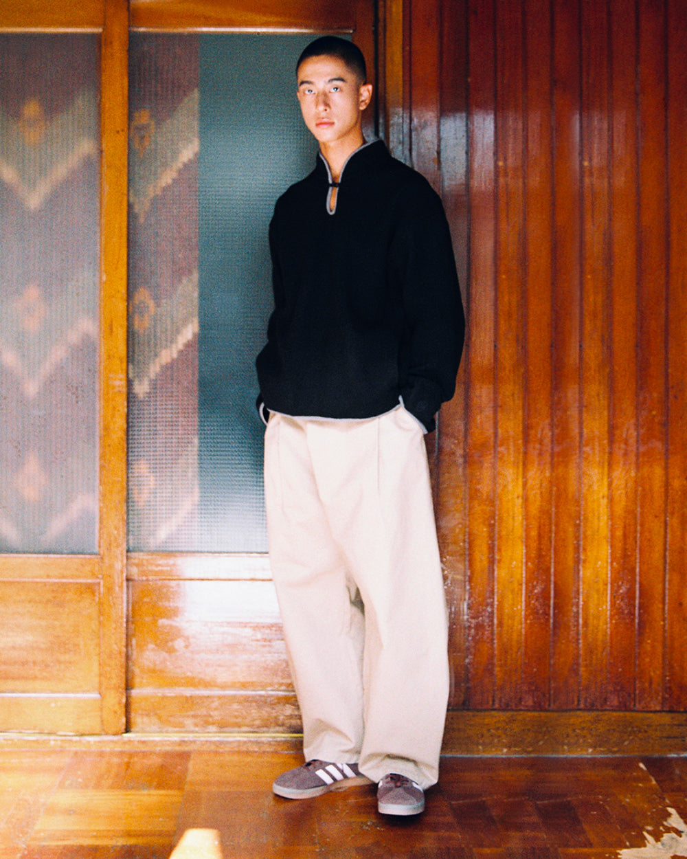 A person stands indoors against wooden sliding doors, wearing a black long-sleeve top layered with the BLACK TEARDROP KNOT FLEECE by Sundae School, beige wide-leg pants, and gray Adidas sneakers, looking at the camera with hands in pockets.