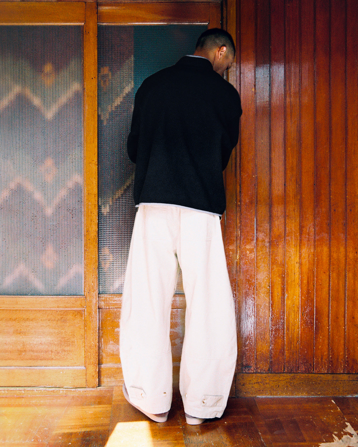 A person with short hair, dressed in a black jacket and wide beige pants, stands with their back to the camera before a wooden wall and door. Sunlight filters across the floor, softly showcasing their cozy BLACK TEARDROP KNOT FLEECE from Sundae School—a subtle nod to East Asian inspired fleece design.