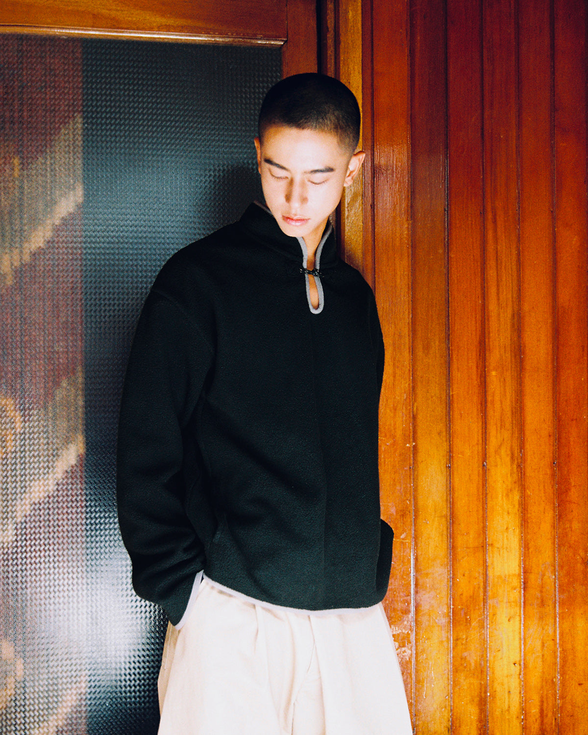 Wearing the Sundae School BLACK TEARDROP KNOT FLEECE zip-up jacket and light-colored pants, a short-haired individual stands with hands in their pockets against a wooden wall, gazing downward.