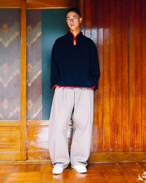 A person stands indoors by wooden paneling, wearing the Sundae School NAVY TEARDROP KNOT FLEECE—a black long-sleeve shirt with red trim and subtle East Asian knotting details—paired with wide beige pants and white shoes, looking directly at the camera.