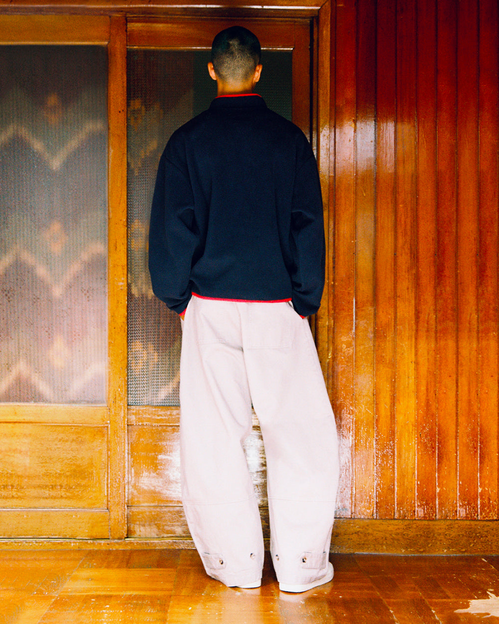 A person with short hair stands indoors, facing a wooden door, wearing the Sundae School NAVY TEARDROP KNOT FLEECE layered under a dark jacket and paired with loose, light-colored pants. The wooden walls and floor create a warm atmosphere that complements the cozy fleece details of their outfit.