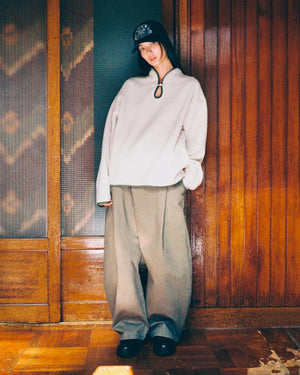 Wearing the TAUPE TEARDROP KNOT FLEECE by Sundae School, along with wide-legged pants, black shoes, and a dark cap, a person leans casually against a wooden wall with a slight smile.