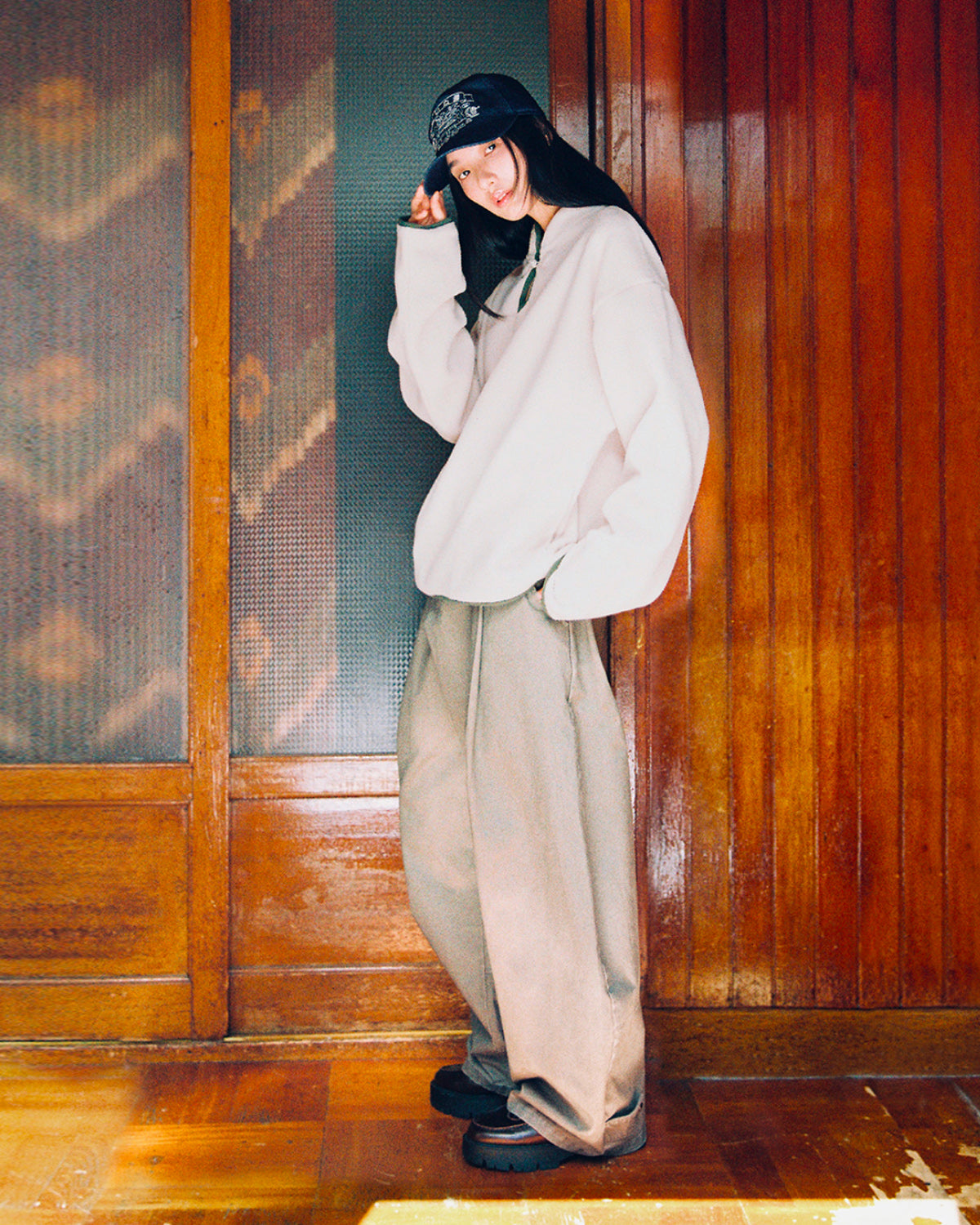 Wearing the TAUPE TEARDROP KNOT FLEECE by Sundae School, along with wide beige pants and a black cap, a person smiles while standing indoors on a wooden floor, one hand holding the brim of their cap.