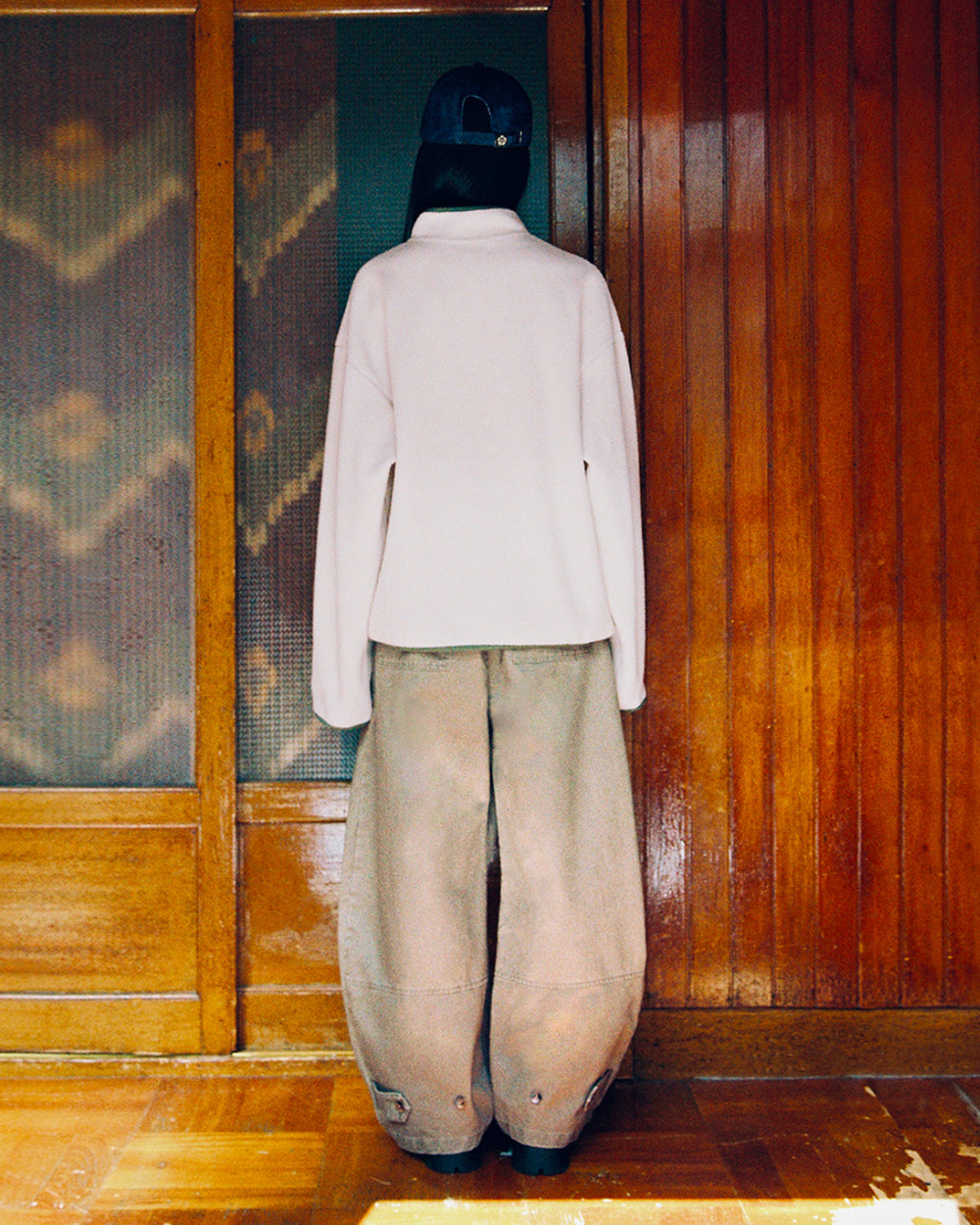 A person stands indoors facing a wooden wall, wearing the Sundae School TAUPE TEARDROP KNOT FLEECE, oversized brown pants, and a dark baseball cap. Their face is not visible.