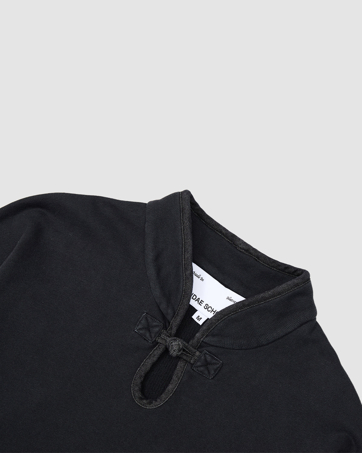 A close-up of the Sundae School BLACK TEARDROP KNOT SWEATSHIRT, featuring a mandarin collar, knotted button closure, and a visible white sizing label inside the collar.
