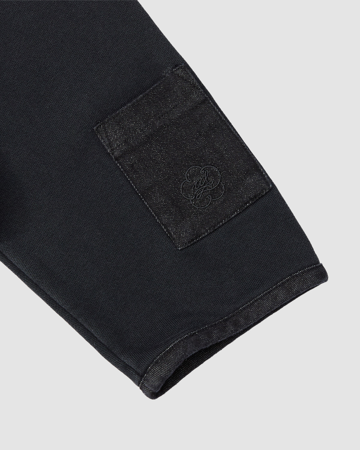 Close-up of the BLACK TEARDROP KNOT SWEATSHIRT by Sundae School: a black cotton sleeve with a dark patch pocket near the cuff, featuring subtle embroidery. Light gray background.
