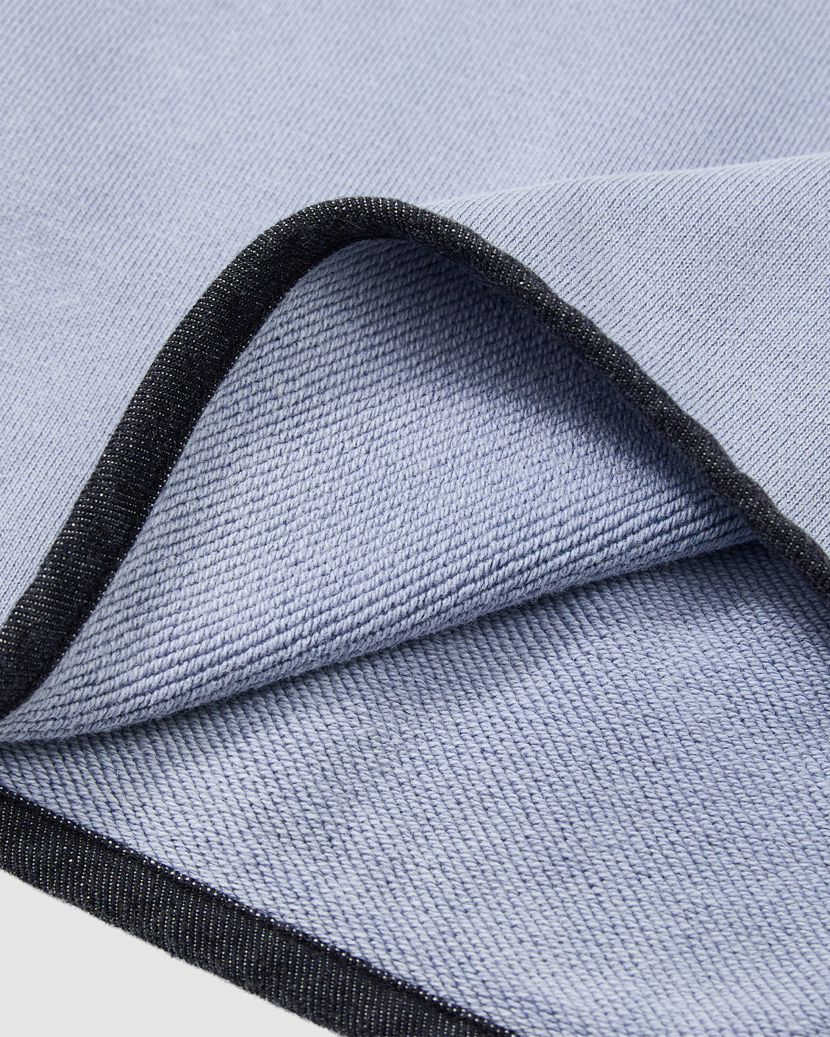 A close-up of Sundae School's SKY BLUE TEARDROP KNOT SWEATSHIRT shows its light blue cotton fabric with a diagonal weave and black denim trim along the edges, with the material elegantly folded.