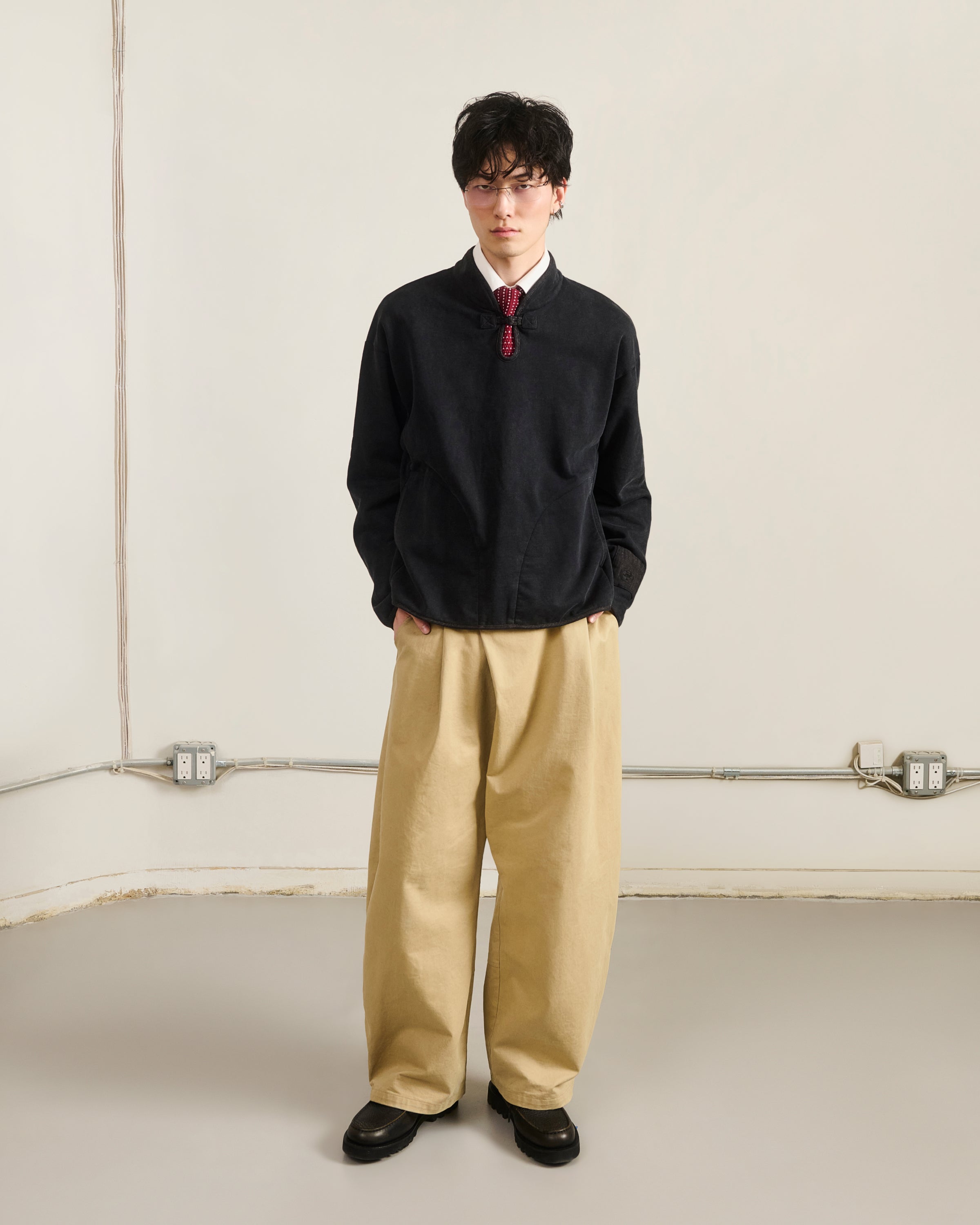 Indoors against a light wall, a person wears Sundae School’s BLACK TEARDROP KNOT SWEATSHIRT with beige wide-leg pants, black shoes, and glasses. Hands in pockets; electrical outlets and cables are visible on the wall.