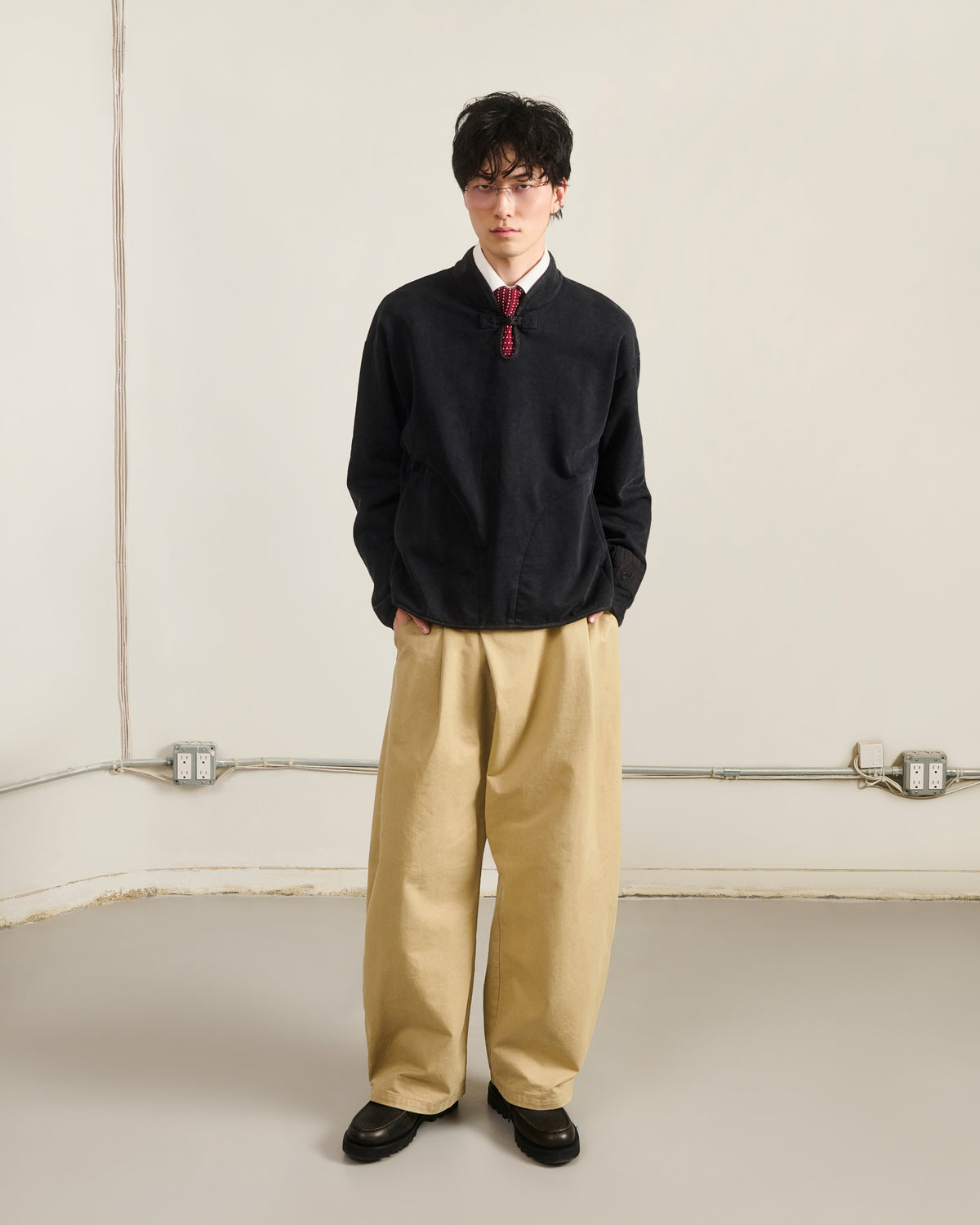 Indoors against a light wall, a person wears Sundae School’s BLACK TEARDROP KNOT SWEATSHIRT with beige wide-leg pants, black shoes, and glasses. Hands in pockets; electrical outlets and cables are visible on the wall.
