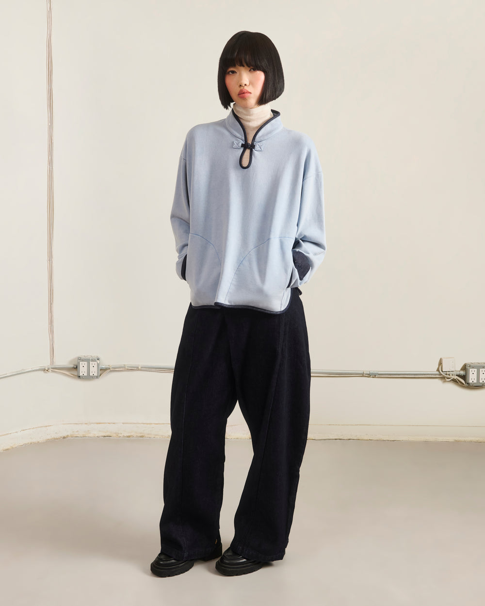 A person with a short black bob stands indoors against a plain wall, wearing the Sundae School SKY BLUE TEARDROP KNOT SWEATSHIRT over a white turtleneck, loose black pants, and black shoes, with hands in their pockets.