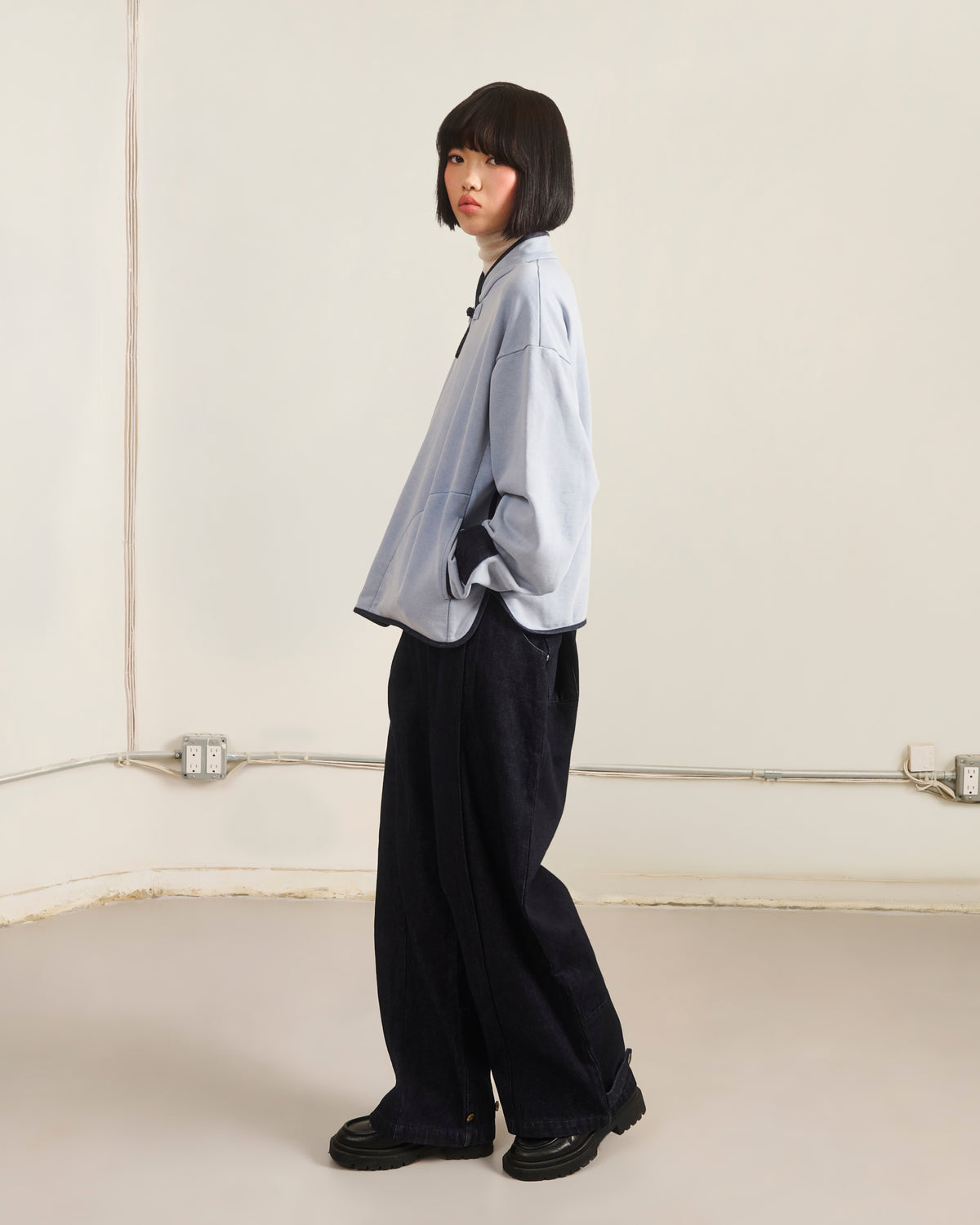 A person with a bob haircut stands sideways in a minimalist room, wearing the Sundae School SKY BLUE TEARDROP KNOT SWEATSHIRT, wide-leg black pants, and black shoes. Plain walls feature visible outlets and cords.