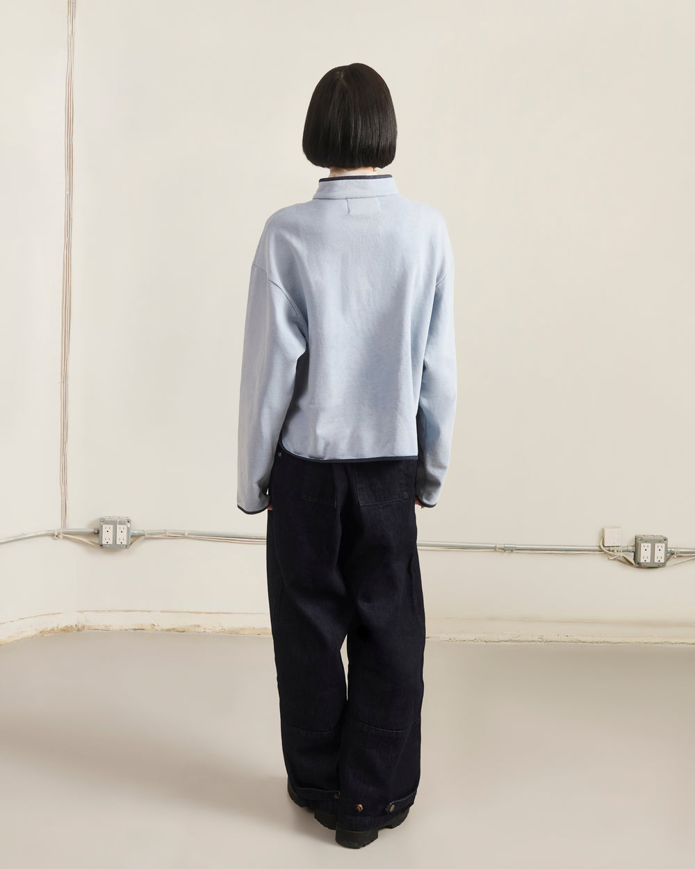 A person with short dark hair, wearing the Sundae School SKY BLUE TEARDROP KNOT SWEATSHIRT and loose black pants, stands facing a plain beige wall with visible electrical outlets and cords near the floor.
