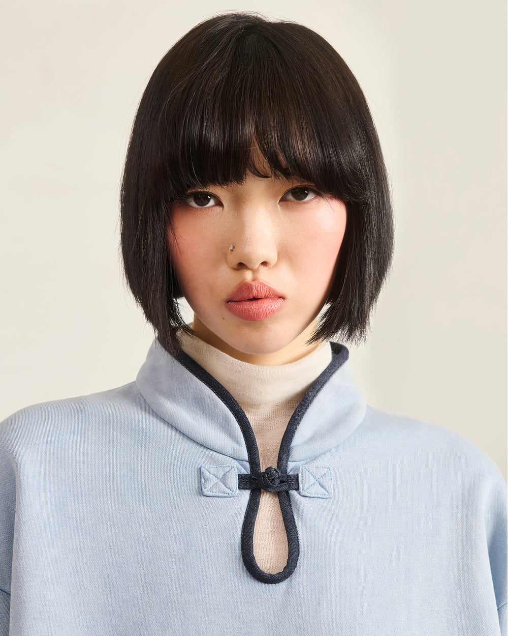 A person with straight, chin-length black hair and bangs wears the SKY BLUE TEARDROP KNOT SWEATSHIRT by Sundae School—crafted from 100% cotton—over a cream turtleneck, staring at the camera with a neutral expression.