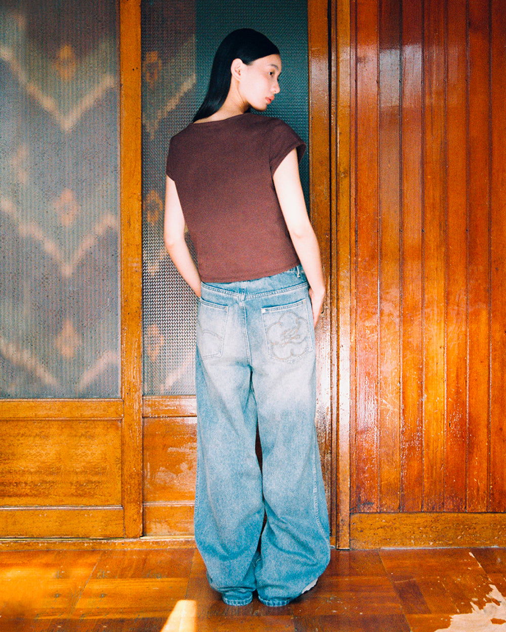 A person with long dark hair, wearing the Sundae School BROWN TIE GORUM BABY TEE and loose blue jeans, stands indoors facing a wooden wall with their hands behind their back.