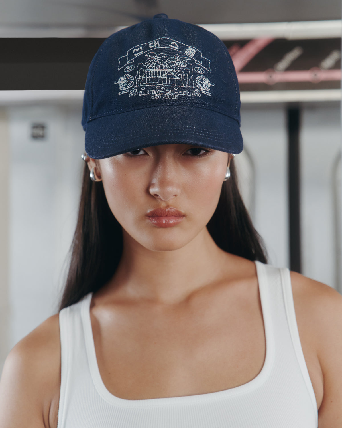 A person with long dark hair wears the Sundae School TIGER DEN CAP—a navy blue denim strapback hat adorned with white Korean folk-art embroidery-inspired graphics—and a white tank top, standing indoors with a neutral expression.