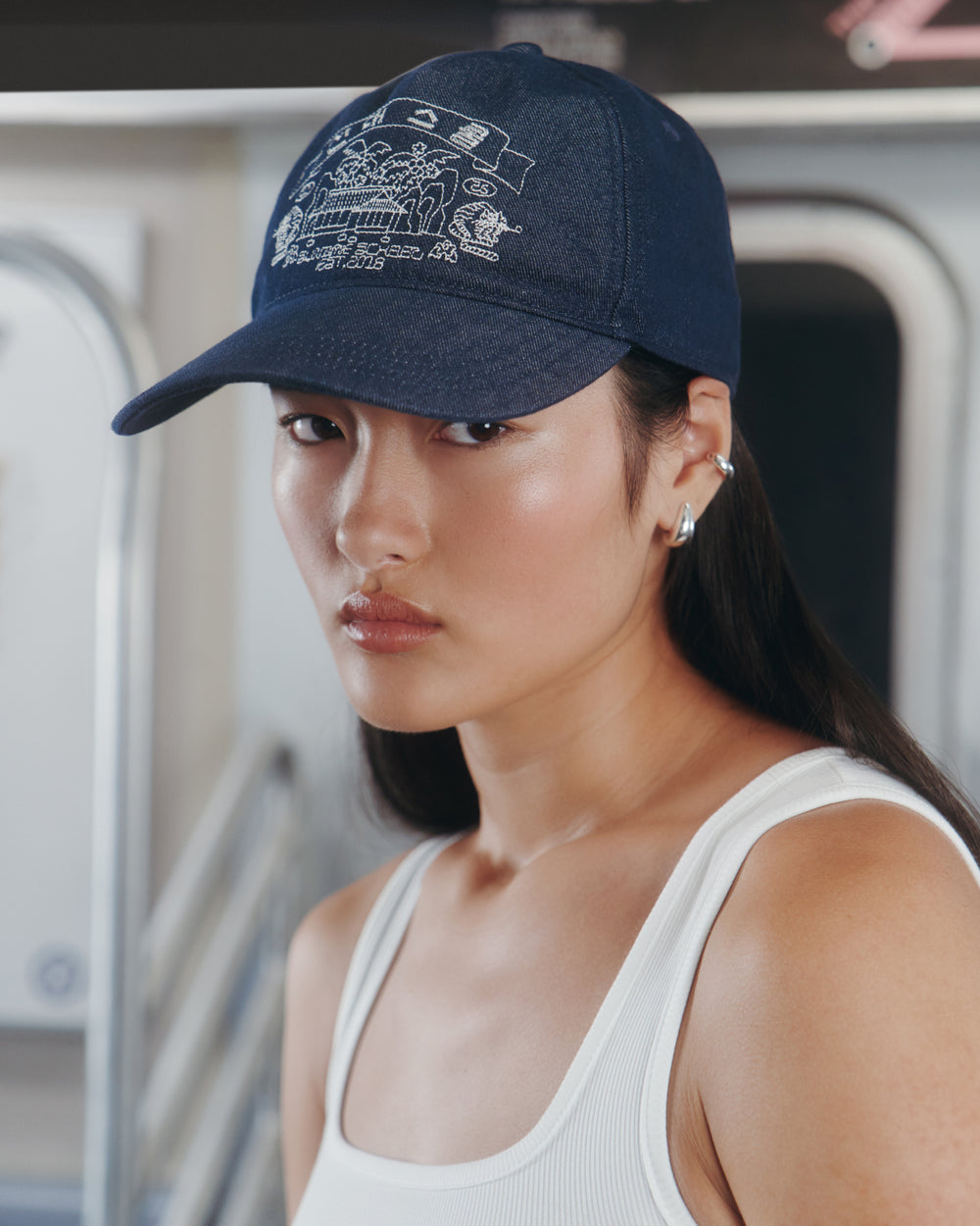A person with long dark hair wears the Sundae School TIGER DEN CAP, a navy denim strapback hat featuring white graphic art, along with a white tank top, while standing in what appears to be a subway train.