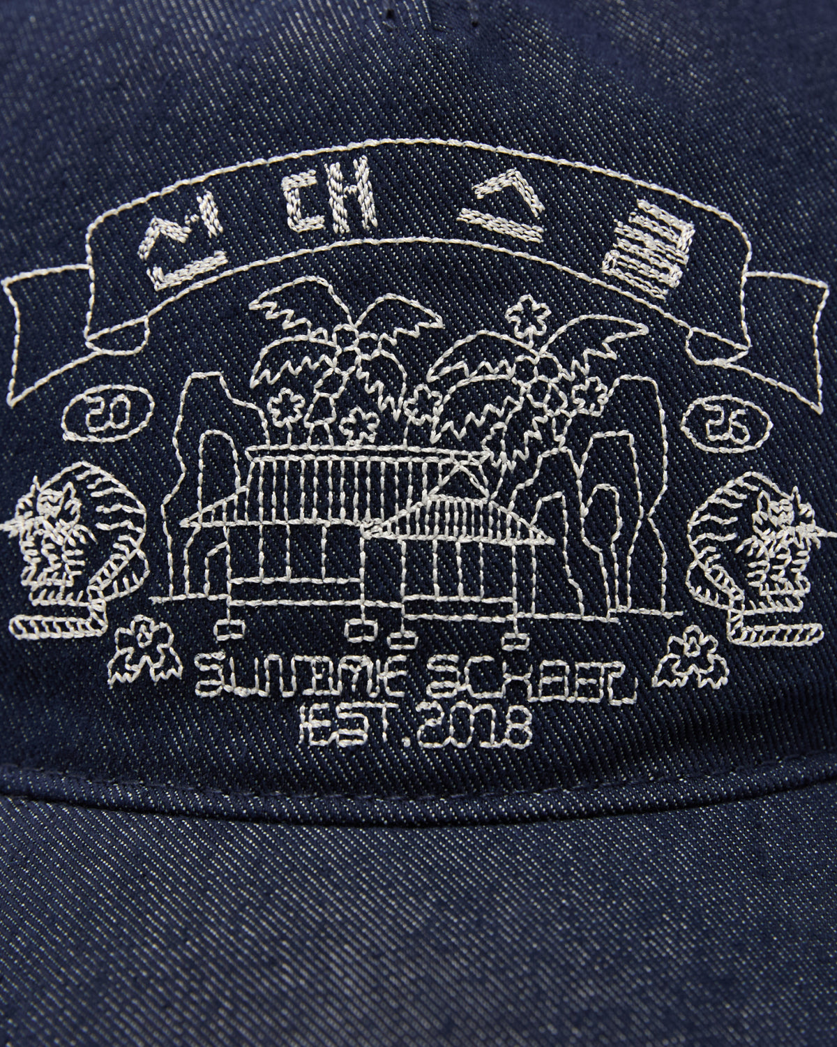 A close-up of Sundae School’s TIGER DEN CAP—a denim strapback hat crafted from a premium cotton blend—shows white Korean folk-art embroidery featuring a building, trees, cliffs, and the text “SUMAME SQUARE EST. 2018,” along with the numbers 20 and 25 and stylized icons above a banner.