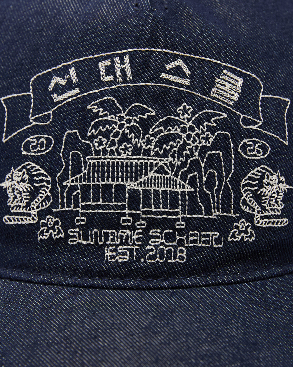 A close-up of Sundae School’s TIGER DEN CAP—a denim strapback hat crafted from a premium cotton blend—shows white Korean folk-art embroidery featuring a building, trees, cliffs, and the text “SUMAME SQUARE EST. 2018,” along with the numbers 20 and 25 and stylized icons above a banner.
