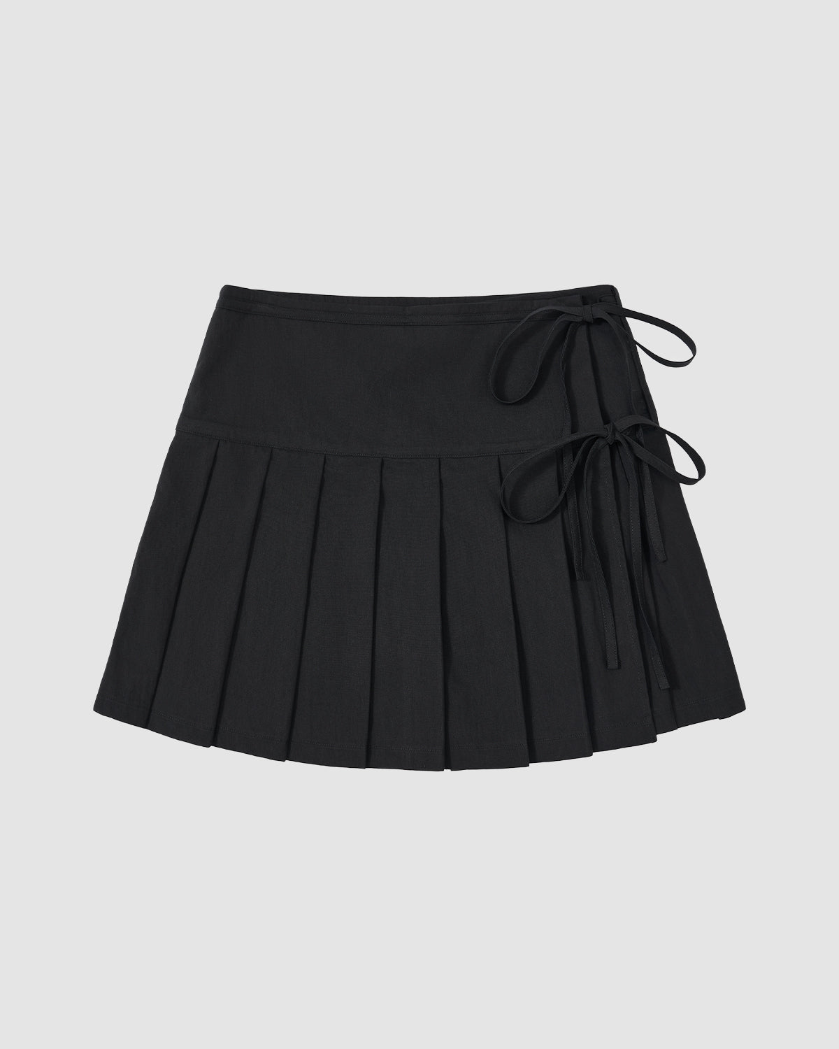 The BLACK UNIFORM APRON SKIRT by Sundae School is a black Korean pleated mini skirt featuring two side tie details on an adjustable waistband, shown against a plain light gray background.
