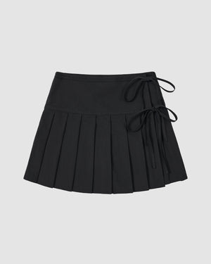The BLACK UNIFORM APRON SKIRT by Sundae School is a black Korean pleated mini skirt featuring two side tie details on an adjustable waistband, shown against a plain light gray background.