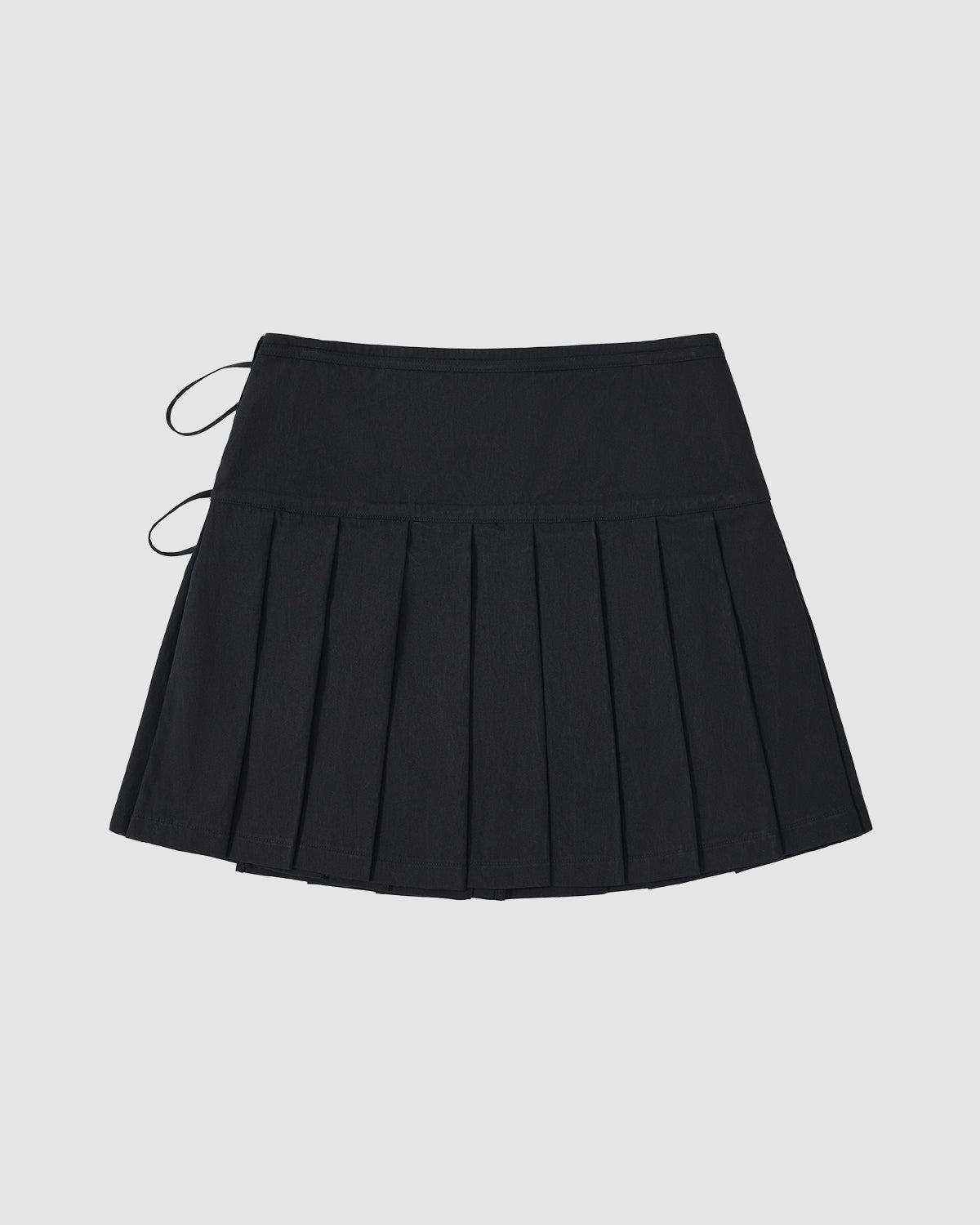 The Sundae School BLACK UNIFORM APRON SKIRT is a black pleated mini skirt with two thin side ties and an adjustable waist, shown flat on a light gray background.